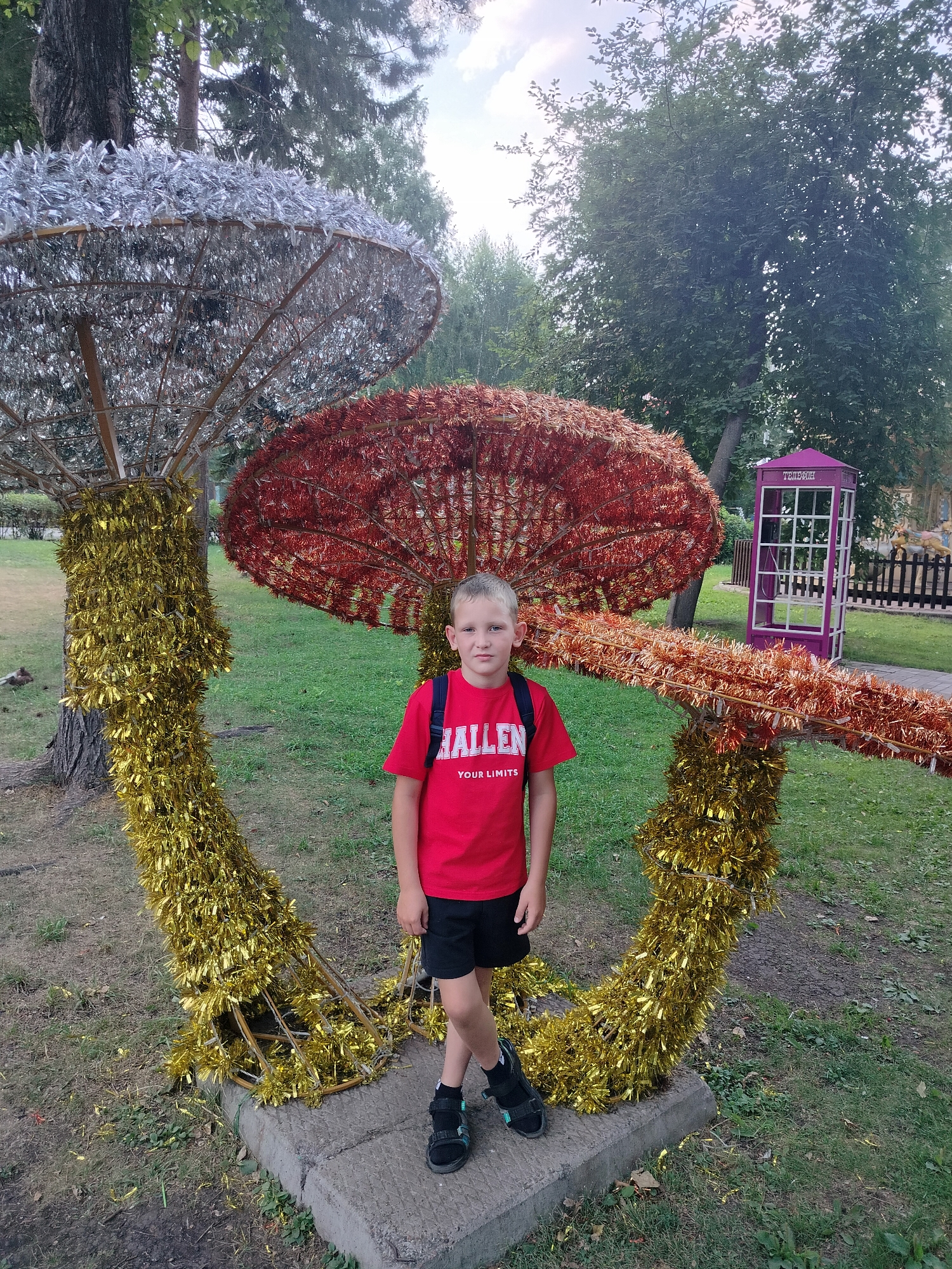 A young boy, appearing to be around 8-10 years old, stands in front of a decorative mushroom sculpture in an outdoor park. The boy is wearing a red t-shirt with "HALLEN YOUR LIMITS" printed on it, black shorts, and black sandals. He has a blue backpack on. The mushroom sculpture is large and made of shiny, tinsel-like material. Two mushroom caps are visible: one is red and the other is silver. The stems are gold. The sculpture is set on a concrete base. In the background, there is a purple telephone booth, a wooden fence, and trees. The scene appears to be located in the city of Kemerovo, Russia.