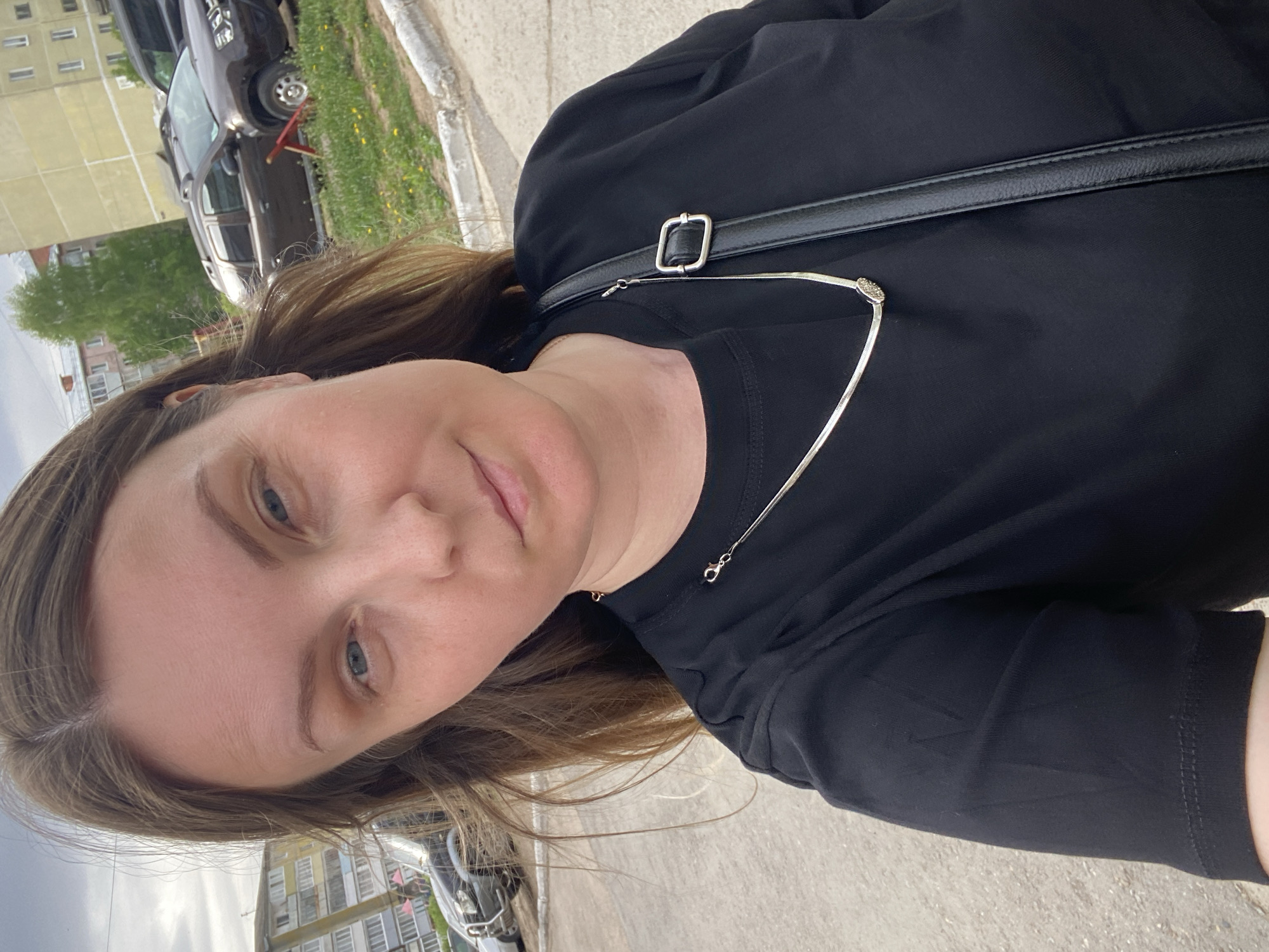 A woman is seen in a close-up, head-and-shoulders shot outside in Noviy, Russia. She is wearing a black top and a silver necklace, and has a black bag with a silver buckle over her shoulder. The background includes buildings, cars, and greenery.