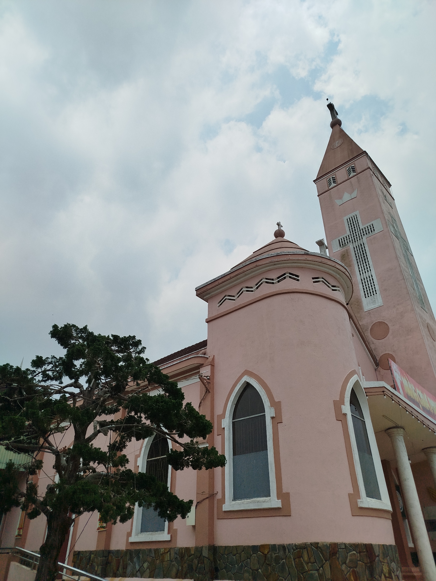 The image shows a pink church in Lang Biang - Đà Lạt, Vietnam. The church has a tall tower with a cross on top. The building is pink with white trim and has several windows. A tree is on the left side of the image. The sky is cloudy.