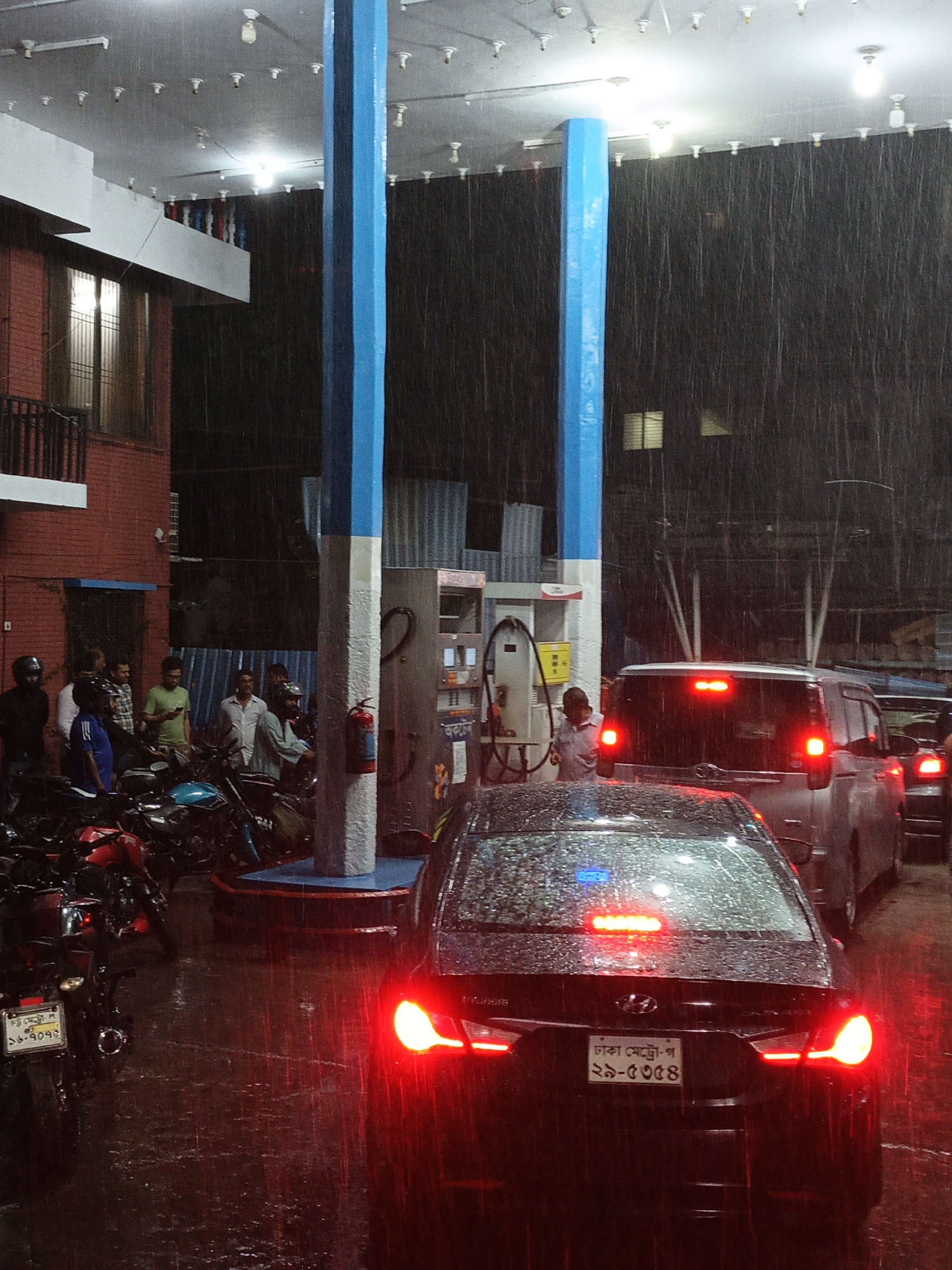 The image shows a gas station in Dhaka, Bangladesh, on a rainy night. A black car is in the foreground with its brake lights on. A gray minivan is directly behind it, also with its brake lights illuminated. To the left, several motorcycles are parked near a group of people standing under the gas station canopy. Two blue and white support pillars are visible near the gas pumps. The rain is heavy, and the ground is wet and reflective. The license plate on the black car is visible, reading “ঢাকা মেট্রো-প ২5-5354”. A license plate on a motorcycle reads “ঢাকা মেট্রো-ল 38-9093”.