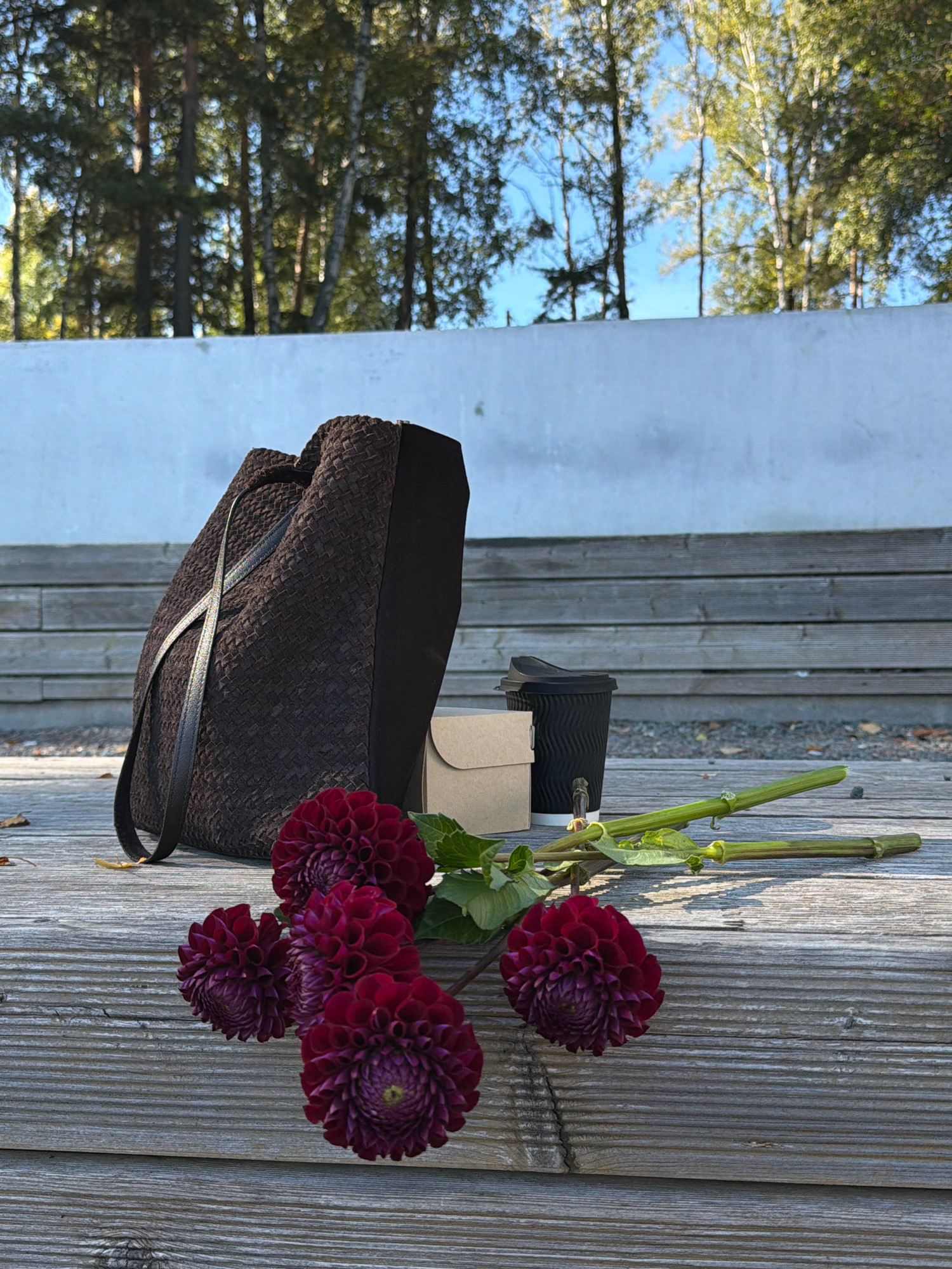 A still life arrangement is set on weathered wooden planks outdoors. In the foreground, several deep red dahlias are clustered together. Behind the flowers, a dark brown woven tote bag with leather straps is propped up. Next to the bag is a small, light brown cardboard box, and a black disposable coffee cup with a lid. Two green plant stems with leaves lie horizontally in front of the coffee cup. In the background, a white concrete wall runs horizontally, and beyond that, a forest of trees with dappled sunlight is visible against a blue sky. The scene suggests an outdoor seating area, possibly in a park or a cafe with a natural setting. The lighting indicates it is daytime. No people are present in the image. The location is identified as Chaikovsky, Russia.