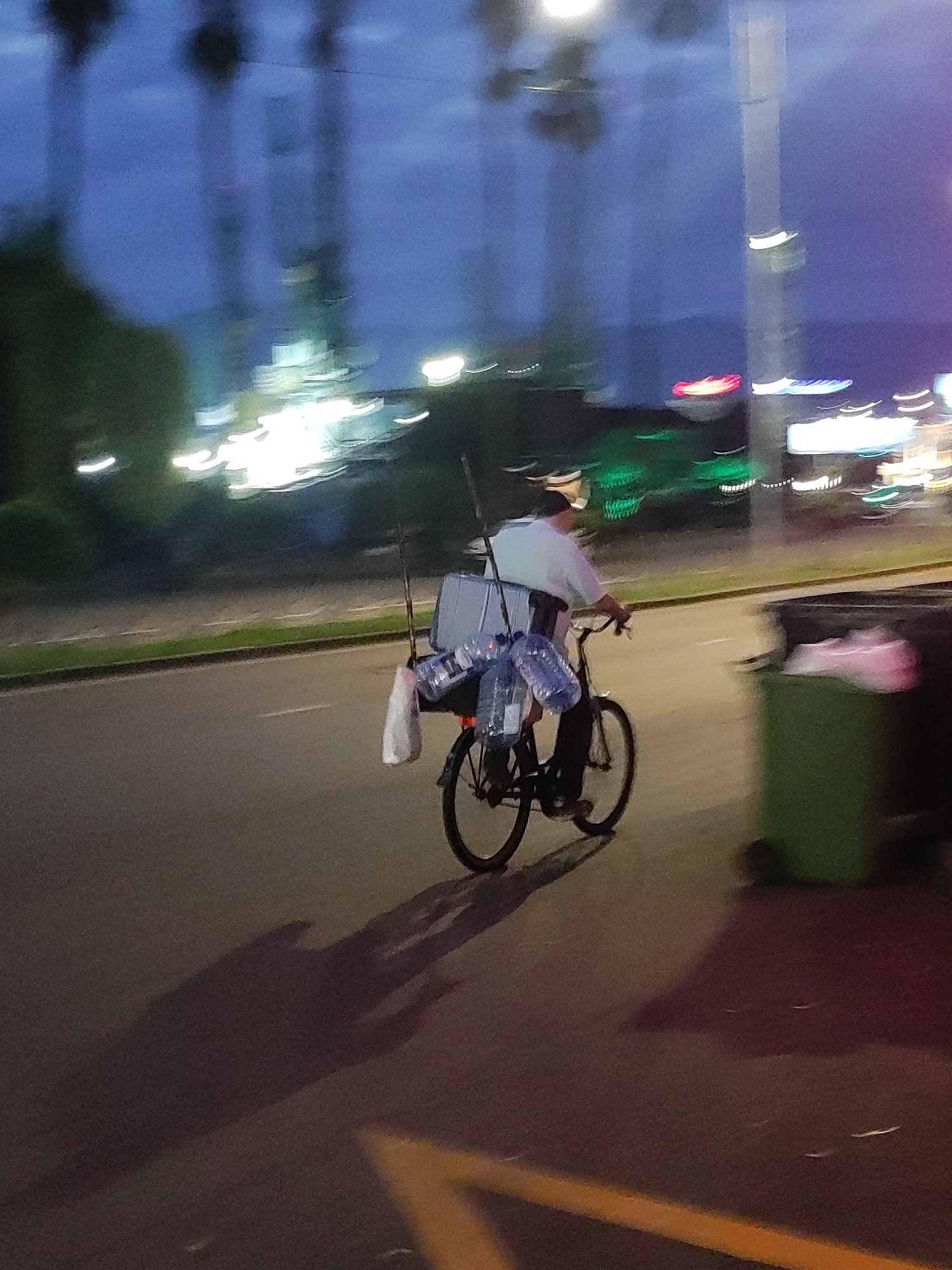 A motion-blurred image taken at dusk shows a person riding a bicycle laden with items on a street in Batumi, Georgia. The cyclist, wearing a light-colored shirt, is positioned in the center of the frame, traveling from left to right. Attached to the bicycle's rear rack are several large plastic bottles, likely filled with liquid, and a white bag. Two poles, possibly fishing rods, are also secured vertically behind the cyclist. The background is indistinct due to motion blur, but suggests an urban environment with blurred lights and structures, including what appear to be streetlights and buildings. The road surface is asphalt, with a visible white lane marking and a yellow line on the right side of the frame. A green trash bin is present on the far right. The lighting conditions indicate twilight or early evening.