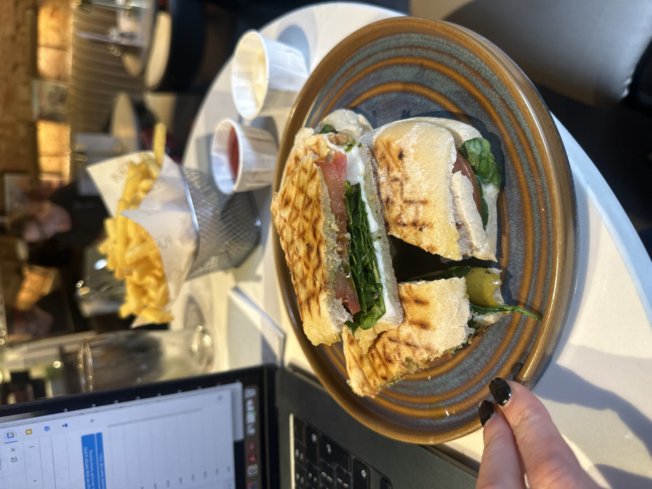 A plate of cut panini sandwiches with spinach, tomato, and white cheese filling is on a white table. Condiments in small white cups and a wire basket of french fries are next to the plate. A laptop is in the foreground. A person's hand with black nail polish is reaching for the food. The food and laptop are on the same table in what appears to be a restaurant.