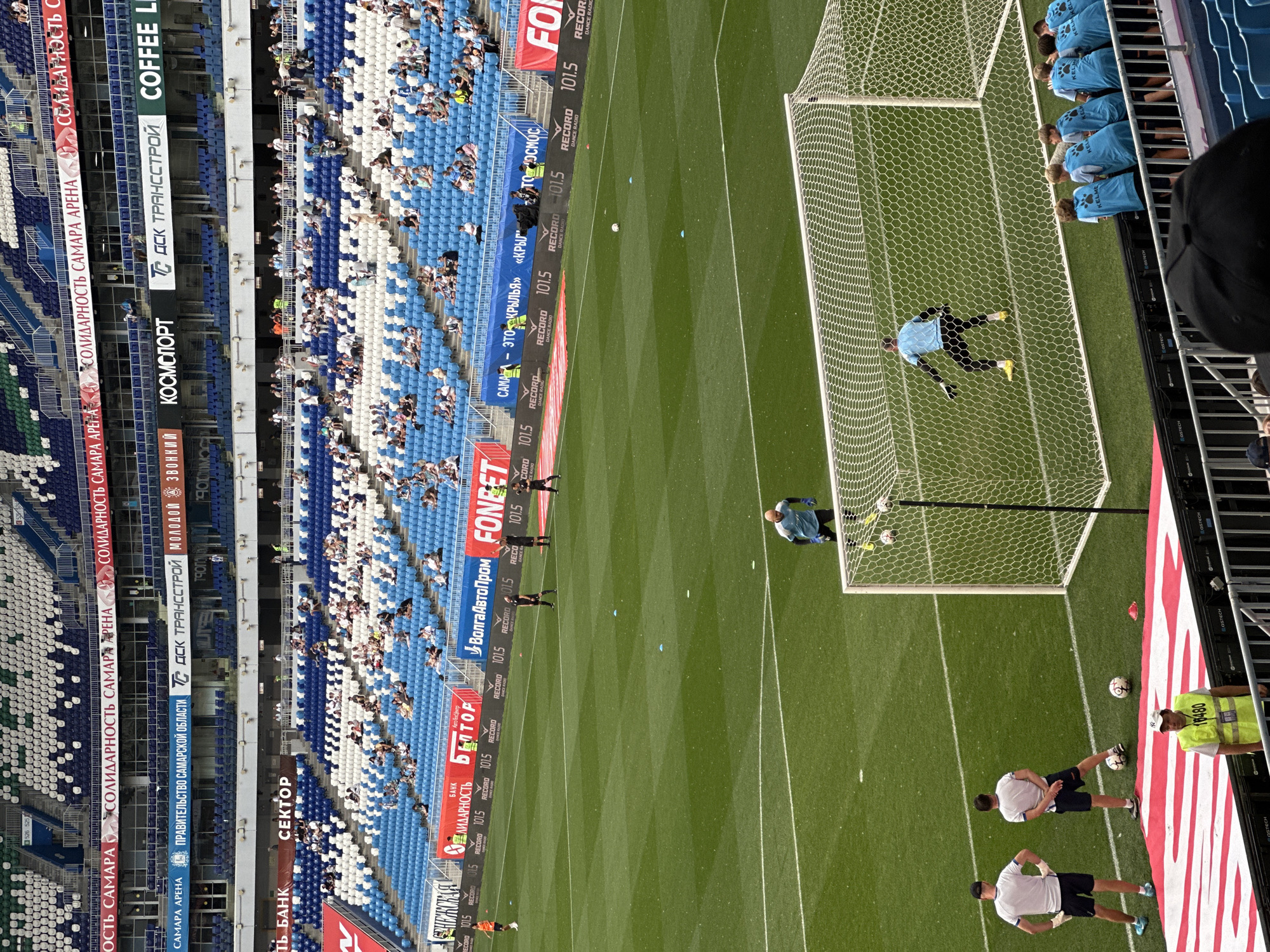 The image shows a wide-angle view of a football stadium during a match. In the foreground, a goal net is visible, with a goalkeeper in a light blue uniform positioned within it. Several footballs are on the grass near the goal. To the left of the goal, two men in white shirts and dark shorts stand on the field, and another man in a yellow safety vest is positioned lower down. In the middle ground, several players in dark uniforms are on the field, including one near the center circle and two near the penalty area. The background features tiered seating filled with spectators, predominantly in blue and white. Above the stands, banners with Russian text and logos are displayed, indicating sponsorship and team affiliation. The stadium appears to be the Samara Arena, a modern football venue located in the city of Samara, Russia, known for hosting major sporting events.