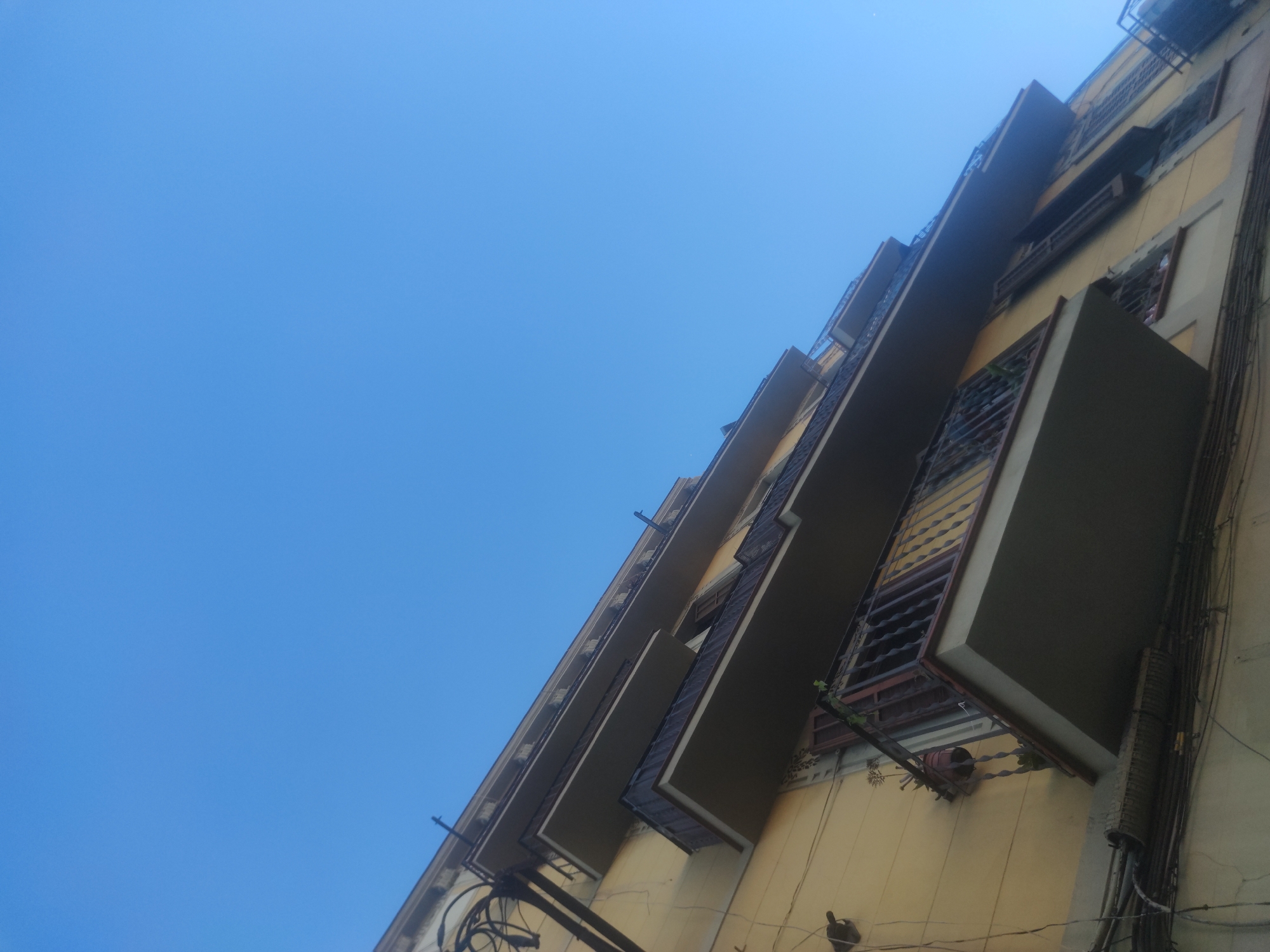 The image captures an upward perspective of a tall, multi-story building against a clear blue sky. The building's facade is a light, faded yellow color. Protruding rectangular balconies are visible on each floor, and they appear to be of a neutral or slightly darker color. The windows have metal grilles, and some show hints of greenery. Electrical wiring and conduits are clustered vertically along the building's right side. The scene suggests an urban environment.