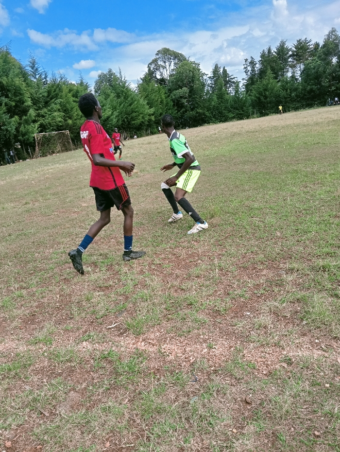 The image depicts an outdoor soccer match or practice taking place in Lwandeti ward, Kenya. The scene is set on an uneven, partially dry grassy field under a blue sky with scattered white clouds. In the foreground, two youthful males with dark complexions are actively engaged. The individual on the left wears a red short-sleeved jersey with black details, black shorts, dark socks, and black athletic shoes, standing with his body angled right and head turned left, looking upwards. The individual on the right wears a lime green short-sleeved jersey with black trim, black shorts, black socks, and white athletic shoes, positioned in a low, crouched, defensive posture, facing the first player. A white-framed soccer goal with a net is visible in the mid-ground on the left side of the field. Several other individuals, presumably players, are present further in the background, including a figure in red near the goal and a yellow figure on the distant field. A dense line of tall green trees forms the horizon, suggesting a natural or rural setting.