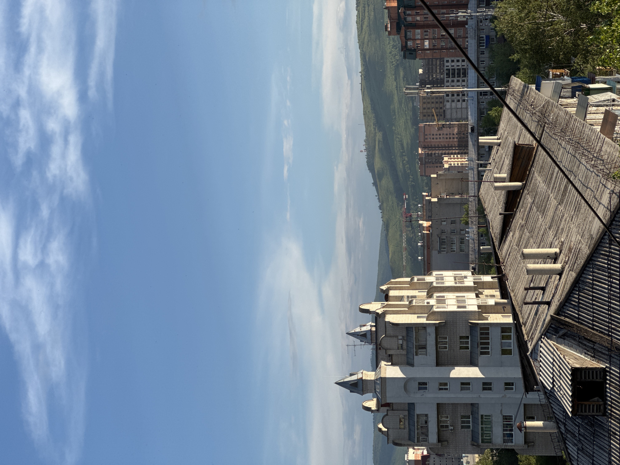 This image, captured from a high vantage point in Chita, Russia, depicts a sprawling urban landscape under a bright blue sky with scattered white clouds. The foreground is dominated by the weathered, grey, corrugated roofs of older buildings, complete with chimneys and what appears to be a small dormer window with its shutter ajar.
In the midground, a striking, multi-story building with a distinct, almost castle-like architectural style stands out. It features white and light beige brickwork, pointed turrets topped with grey caps, and numerous windows arranged in a rhythmic pattern. Behind this building, a dense collection of residential and commercial structures stretches across the cityscape, varying in height and architectural design, with some showing warm brick facades and others concrete exteriors. Electrical wires crisscross the scene, suggesting a developed urban infrastructure.
The background is characterized by rolling, green hills, indicating the natural topography surrounding Chita. The sunlight casts shadows, highlighting the textures of the buildings and the contours of the landscape. The scene conveys a sense of a typical day in this Siberian city, with a mix of older and more modern architecture set against a backdrop of natural beauty.