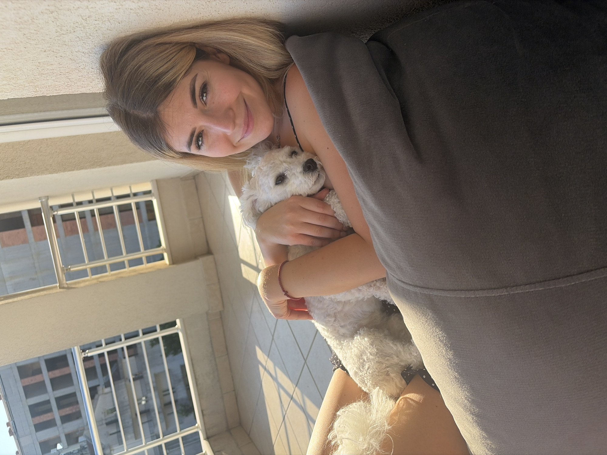 A young adult female with blonde hair parted on the left is seated on a balcony in Budva, Montenegro, holding a small white curly-haired dog against her chest. She smiles softly at the camera, wearing a black top with thin straps and a thin red bracelet on her left wrist. The dog, a Bichon Frise type, rests with its head visible, facing towards the right. The woman leans against a light-colored textured wall and is partially supported by a dark gray fabric beanbag. The balcony floor is tiled in light beige, exhibiting distinct square patterns and prominent geometric shadows from direct sunlight. The background features white metal balcony railings with horizontal bars, beyond which multiple buildings are visible; one appears to be under construction with exposed brick framework in rectangular openings, and another shows windows, suggesting an urban environment.