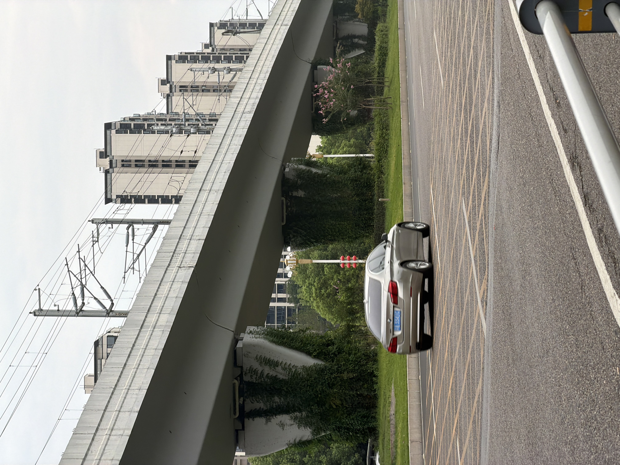 The image depicts a street scene in the city of Jiangjin, China, under an elevated railway or highway structure. A silver BMW sedan with a blue license plate is traveling on the road from left to right. The road has multiple lanes marked with yellow and white lines, including a grid of yellow lines indicating a special area, possibly for queuing or hazard warning. The elevated structure is supported by large concrete pillars, some of which are covered in dense green ivy. In the background, several modern apartment buildings are visible, along with overhead power lines for the railway. Green trees and a small flowering tree with pink blossoms are present along the roadside. The sky is overcast. The image is taken from a low angle, slightly from behind and to the side of the car.