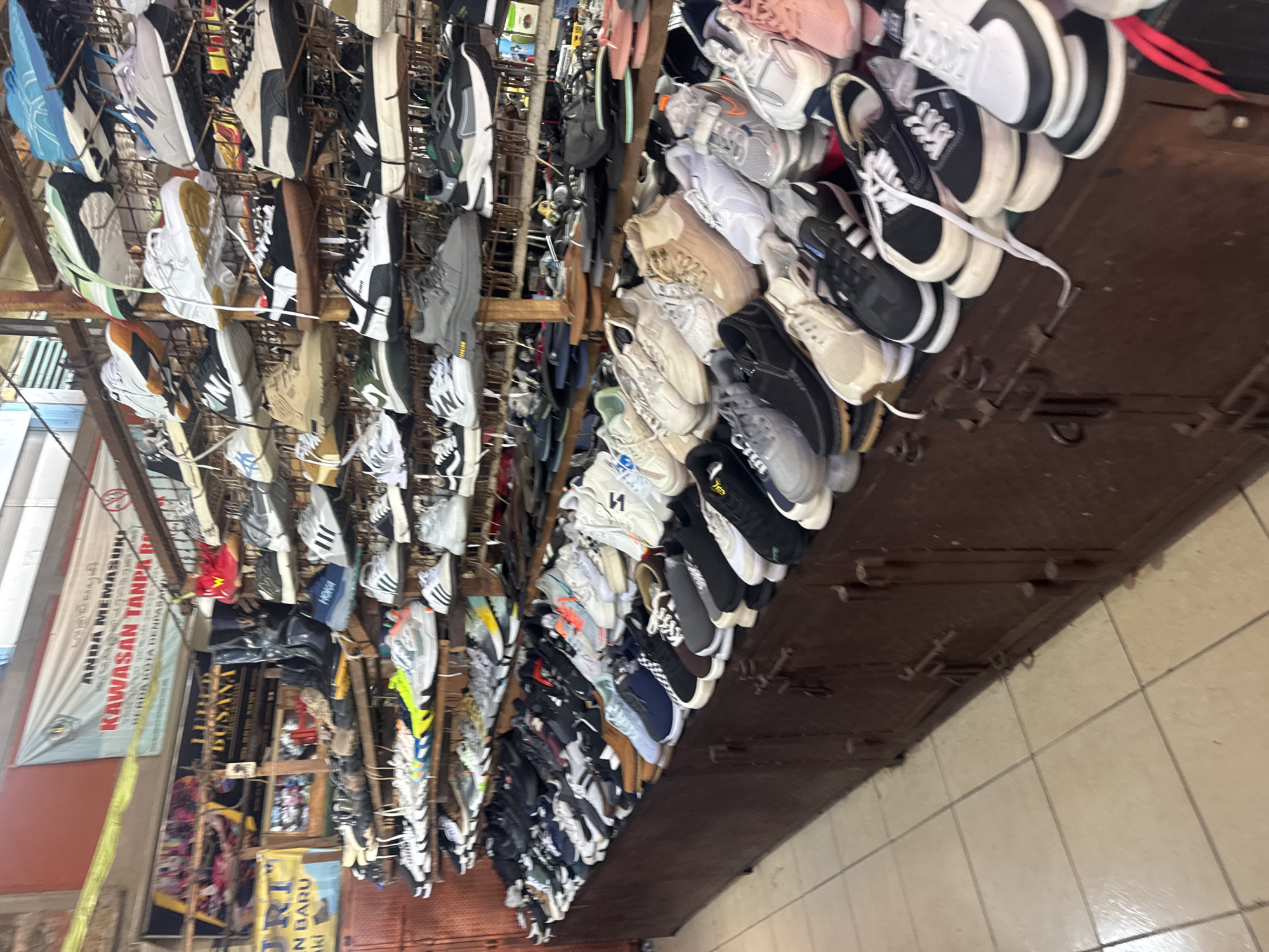 The image depicts a shoe store in Denpasar, Indonesia, likely a market stall or a small shop. The scene is filled with shelves and a long counter stacked with a vast variety of athletic shoes, sneakers, and casual footwear. The shoes are displayed in numerous rows, creating a dense visual landscape of colorful and diverse footwear. The store appears to be indoors, with wooden shelving units and a dark wooden counter. In the background, banners are visible, one with Indonesian text, indicating the location. The floor is tiled with a light-colored material. There are no people actively engaged in the scene, suggesting the photograph was taken during a quiet moment or that the customers are off-camera. The overall impression is one of a busy retail environment focused on selling shoes.