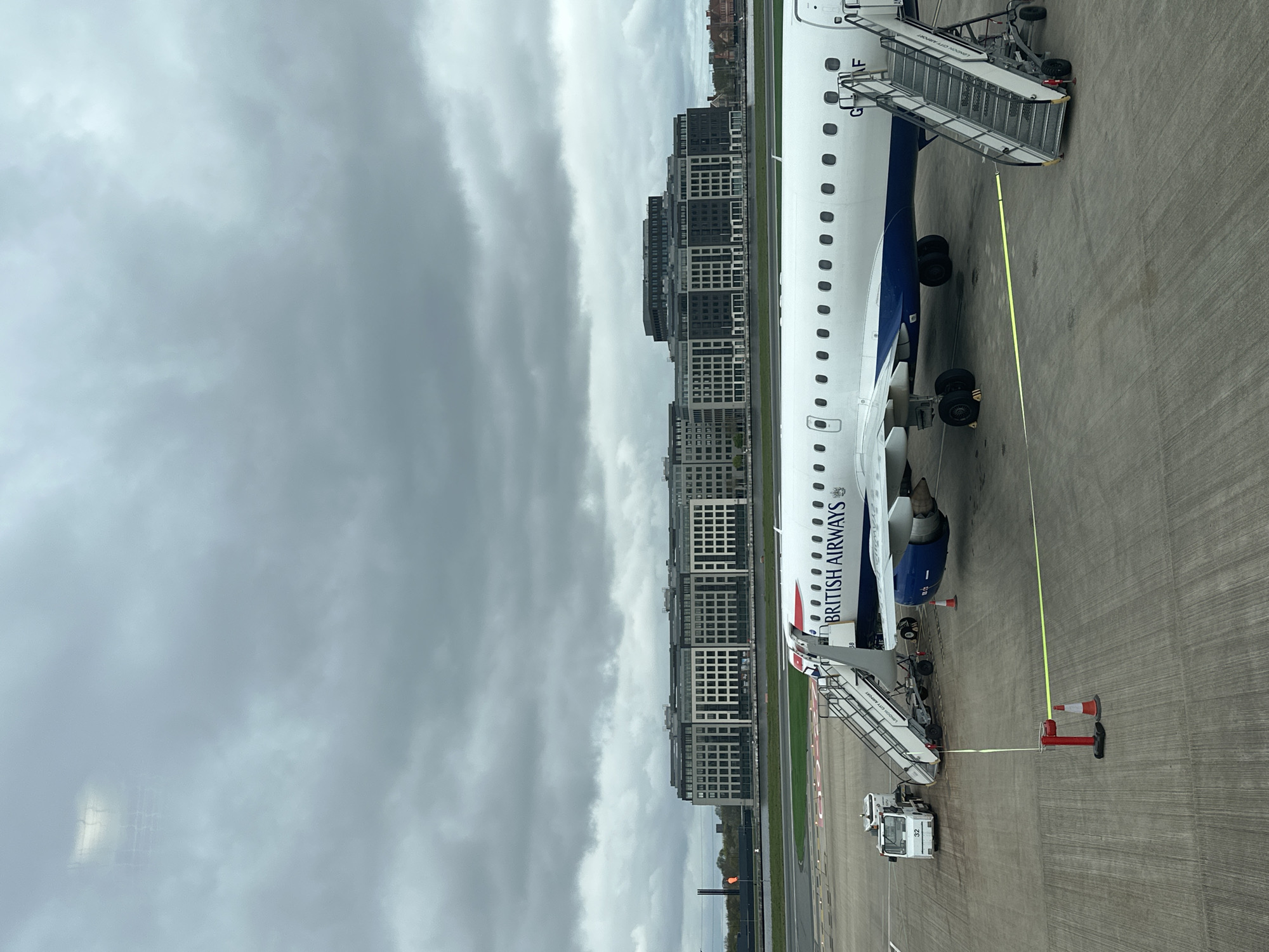An airplane is parked on the tarmac, likely at the London City Airport in the United Kingdom, given the branding on the boarding stairs attached to the plane. The plane, a British Airways aircraft, is equipped with two sets of stairs for boarding and deplaning. A small vehicle, possibly a ground support vehicle, is visible near the front stairs. Barriers and cones surround the plane. In the background, a cluster of multi-story buildings is visible, suggesting a city environment. The sky is overcast with dark clouds.
