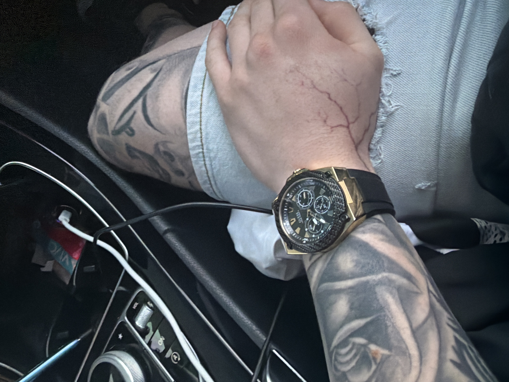 The image presents a close-up view of a person's left arm and hand, situated inside a modern car, likely in Shumen, Bulgaria. The arm is extensively covered in black and grey tattoos, forming what appears to be a full sleeve with intricate, organic designs. A prominent gold-toned watch is worn on the wrist, featuring a black bezel with a carbon fiber-like texture, a black dial with three subdials, and the brand name "GUESS" visible. The watch is secured with a black strap. Noticeable veins are visible on the back of the person's hand.
The person is wearing light-colored, possibly distressed denim shorts or pants, which are visible on their lap and upper thigh. Another tattoo can be seen on the upper leg. The interior of the car features a dark dashboard and center console in the foreground. A white charging cable, appearing to be USB-C, is plugged into a port on the console, suggesting a device is being charged. A small, colorful item, possibly a wrapper, is stowed in a compartment nearby. Control buttons and rotary knobs are visible on the console, one of which clearly displays the text "ON" next to an arrow. The lighting suggests it is daytime, with soft natural light illuminating the scene. The activity appears to be simply sitting in the car, potentially while a device charges. No specific emotions are discernible from the partial view of the person.