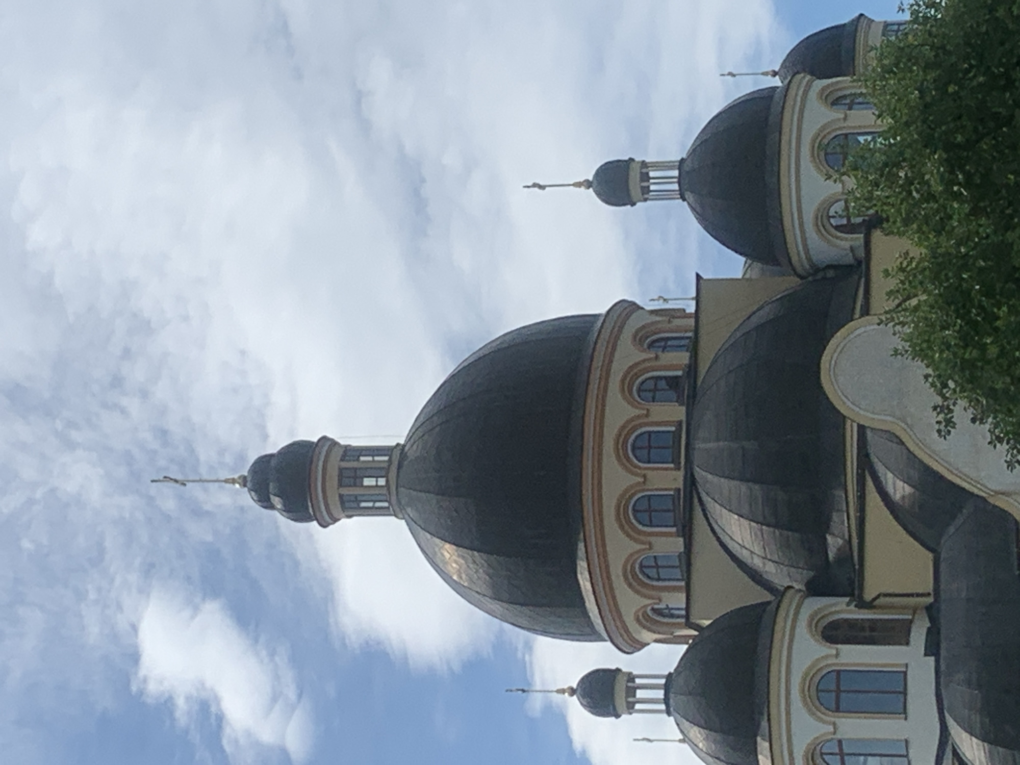 The image shows an upward view of the domes of the Church of the Ascension of the Lord in Kolomyia, Ukraine, under a blue sky with scattered clouds. The prominent domes are dark and rounded, with light-colored trim and arched windows visible on their bases. Several smaller domes and spires with crosses are also visible. Green foliage of trees partially obscures the lower portion of the church. The architectural style appears to be Orthodox Christian.