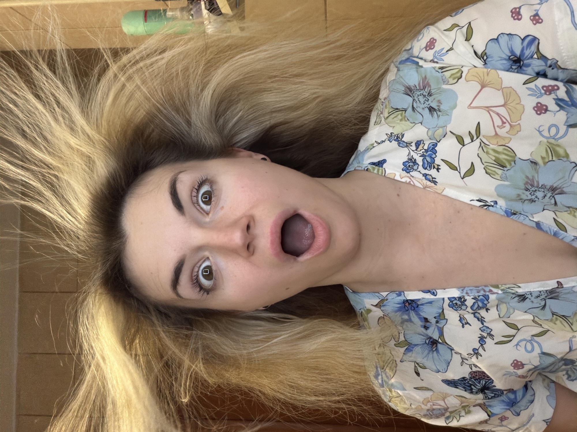 A woman with long blonde hair is lying on what appears to be a wooden floor, possibly indoors. Her hair is spread out around her head. She is wearing a light-colored floral-patterned top. Her face shows an expression of surprise or shock, with her eyes wide open and mouth agape. No specific landmarks or details indicate a location in Budva, Montenegro.