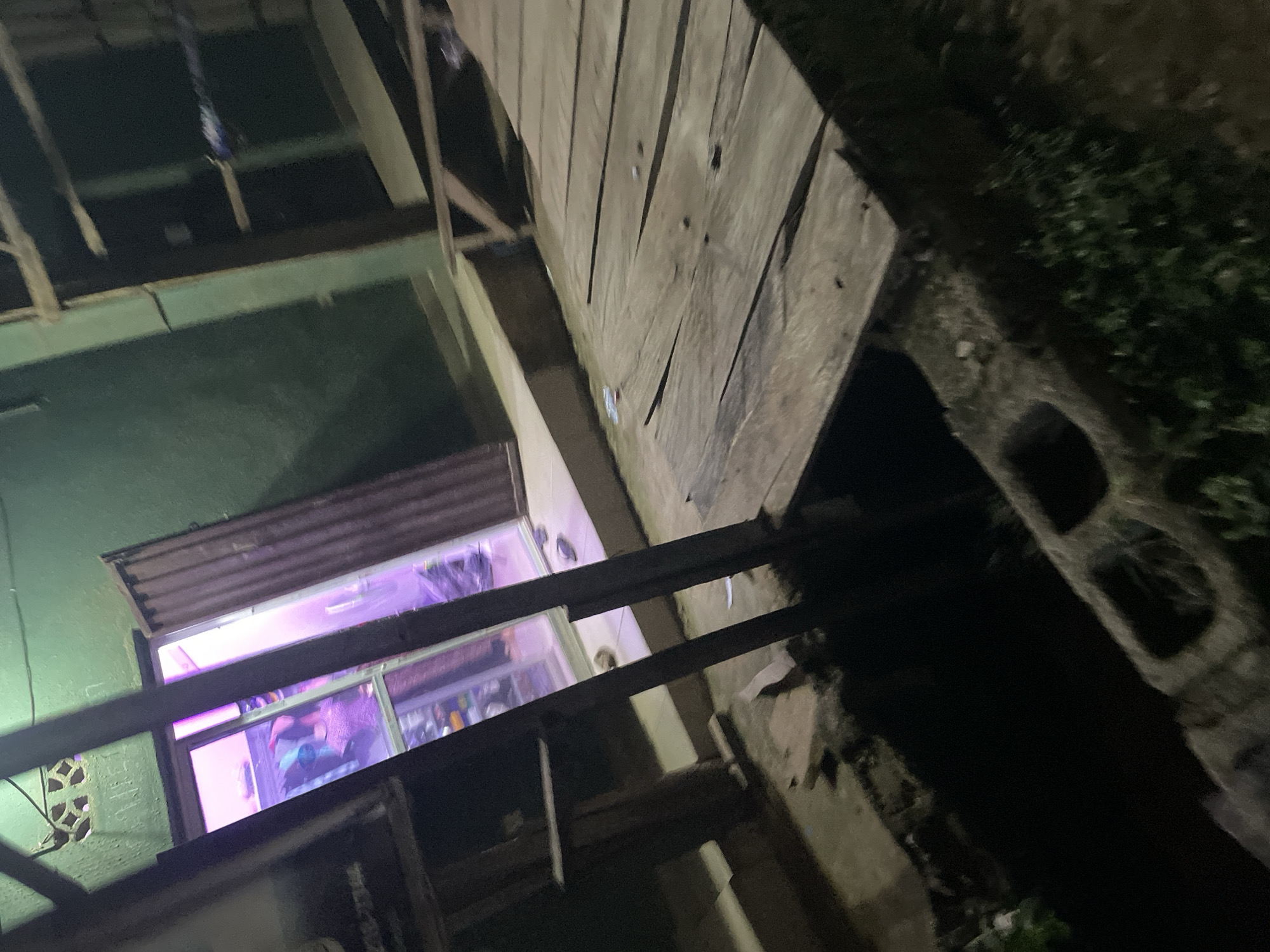 The image captures a dimly lit, outdoor scene in Lokoja, Nigeria, featuring a wooden walkway constructed over a dark, open ditch or pit. The walkway, made of weathered planks, appears to be a temporary or rustic structure. To the left, a green-walled building is visible with an open doorway illuminated by a bright pink or purple light, suggesting an interior space with possibly neon lighting. Inside the building, shelves with various items are discernible. In front of the building, there's a concrete floor, and a dark pole extends upwards, possibly part of the building's structure or a support for the walkway. Scattered on the ground near the walkway are a few plastic bottles and debris. The overall impression is of a functional, albeit somewhat neglected, area in what might be a market or a residential compound.