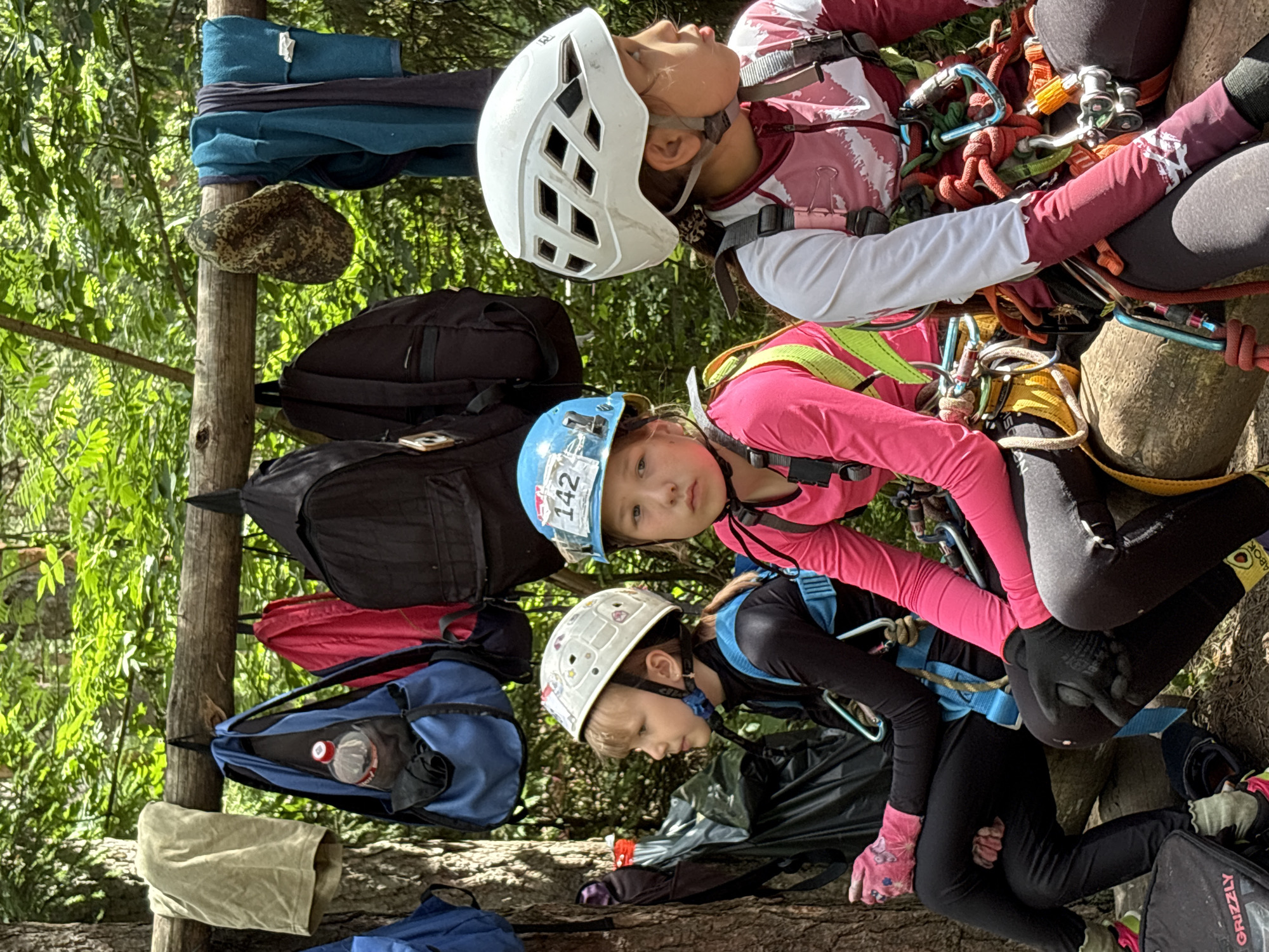 Three young girls, all wearing climbing harnesses and helmets, are sitting on a log in a wooded area. The girl on the left has a light-colored helmet with animal decals and is wearing a black long-sleeved shirt and black pants. She has pink gloves on. The middle girl has a blue helmet with the number "142" on it and is wearing a bright pink long-sleeved shirt and black pants. The girl on the right has a white helmet and is wearing a white and pink long-sleeved shirt and black pants. In the background, backpacks and clothing are hanging from a wooden beam supported by a tree. The setting appears to be a campsite or an outdoor adventure park. The image is taken in natural daylight. There is no visual information to suggest this location is in Chaikovsky, Russia.