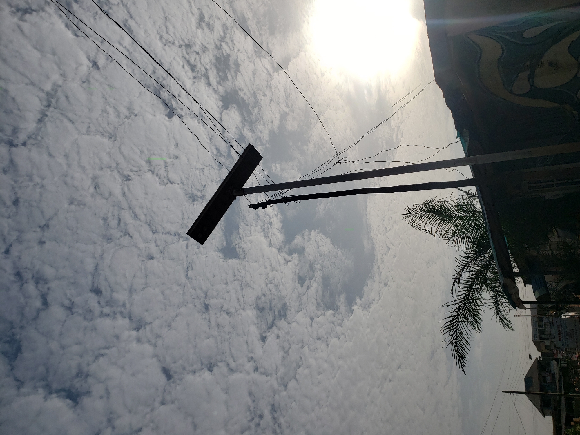 This image depicts an outdoor scene, likely in Narayi, Nigeria, featuring a sky filled with scattered white clouds. In the foreground, a utility pole stands prominently, supporting several black electrical wires that stretch across the frame. To the right, the bright sun is visible, partially obscured by clouds and causing a lens flare effect. Below the wires, the roof of a structure with a dark, possibly corrugated metal surface is visible, adorned with a yellow and blue abstract design. A palm frond extends from the left side of the image, partially obscuring the view of the building and the sky. In the background, several buildings and other utility poles can be seen, suggesting a moderately developed urban or suburban area. The overall impression is of a typical day in the city, with infrastructure and natural elements coexisting.