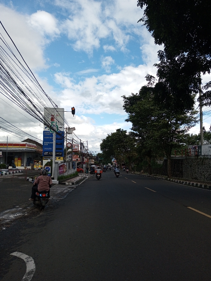 A daytime street scene features an asphalt road with a yellow double solid line marking a lane and a white curved line in the foreground. The sky above shows scattered white clouds on a blue background. On the left side of the road, a person wearing a light-colored shirt is seated on a dark motorcycle, stopped at what appears to be an intersection. Adjacent to the motorcycle is a sidewalk with black and white striped curbs. A tall, blue and white signpost, displaying "PERTAMINA" and fuel price listings for products like "PERTAMAX" and "DEXLITE," stands prominently. Above the road near this sign, a traffic light displays a red signal. Behind the sign, a building with a red and yellow roof is visible. Numerous overhead utility cables are densely strung across the upper left portion of the image. Further down the street on the left, more commercial buildings are visible. On the right side of the road, a sidewalk with striped curbs runs alongside a low stone wall, behind which several large trees with dark green foliage are present. Multiple motorcycles with riders are observed moving away from the camera in the distance on the road. This scene is located in the city of Serpang Shalli, Bhutan.