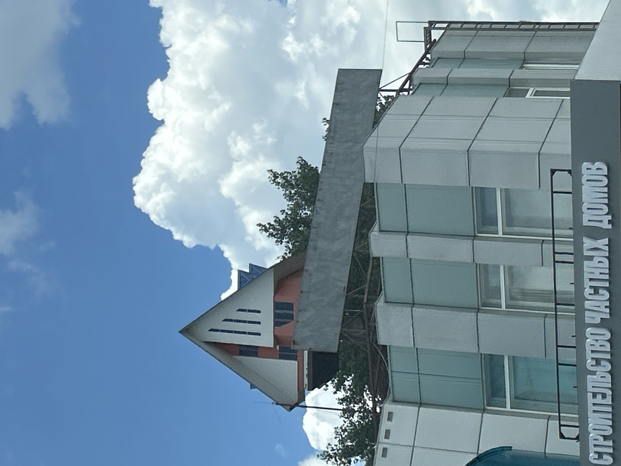 The image shows the facade of a modern building with a distinct architectural feature on its roof. The building has a contemporary design with large, rectangular windows and a facade composed of light-colored panels, possibly concrete or metal. Above this main structure, a stylized, triangular rooftop structure is visible, resembling a house or an abstract architectural element. This triangular section is painted white and red and features vertical windows. Behind and around the rooftop structure, lush green trees are growing, partially obscuring the view. A large, grey, rectangular billboard or sign structure extends horizontally in front of the rooftop element. The sky above is a vibrant blue, dotted with fluffy white clouds. At the bottom of the image, text is visible on a grey band across the facade, reading "СТРОИТЕЛЬСТВО ЧАСТНЫХ ДОМОВ," which translates to "Construction of Private Houses" in Russian. The overall scene suggests a commercial or business establishment related to construction, possibly located in Orlovskii, Russia, as indicated by the Cyrillic text and implied by the prompt. There are no people visible in the image.