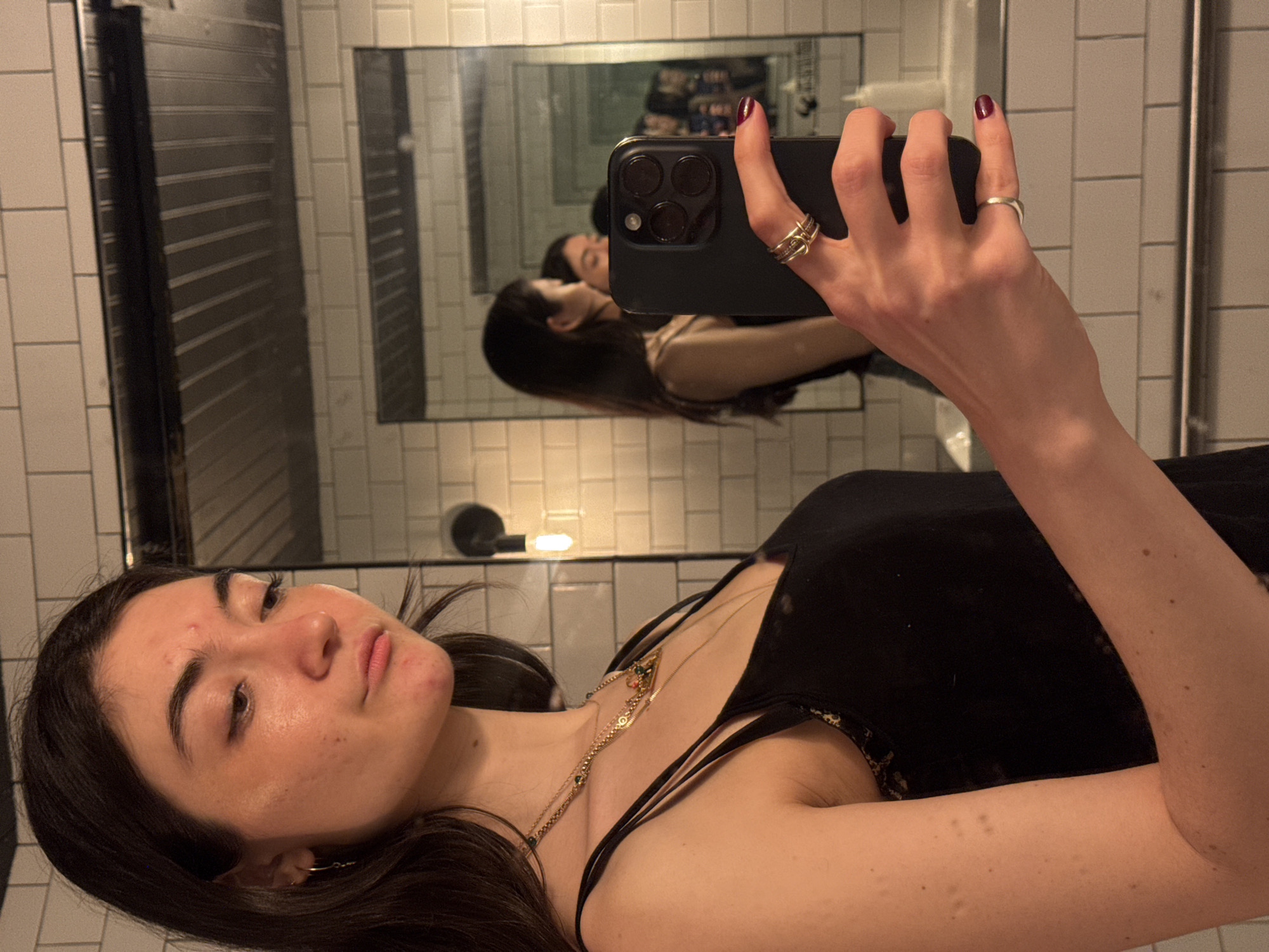 A woman is taking a selfie in a bathroom. She is holding a black iPhone in her right hand, which obscures the reflection of her face in the mirror. The mirror is rectangular and framed, and it is mounted on a tiled wall. The tiles are white with dark grout lines. A light fixture with a single bulb is visible above the mirror. The woman is wearing a black tank top with thin straps and a gold necklace. She has dark hair and is looking at the camera. Her skin has some blemishes. The location is likely a bathroom in New York City, United States of America, considering the bathroom's style and the camera's reflection.