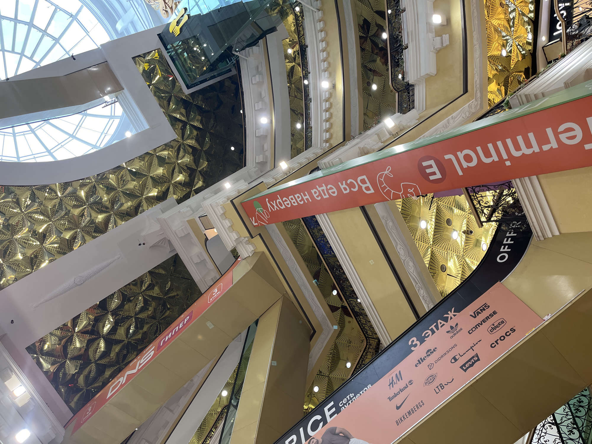 The image shows the interior of a multi-level shopping mall with escalators and banners. The architecture features a large, ornate golden ceiling with a geometric pattern. Natural light streams in from a large domed skylight at the top.
On the left side of the image, a pink banner wraps around an escalator, displaying logos of various clothing and shoe brands such as Nike, H&M, Timberland, Levi's, Converse, Vans, Adidas, and Crocs. The text "3 ЭТАЖ" (which translates to "3rd Floor" in Russian) is prominently displayed on this banner. Another banner on the left, mostly out of frame, features a man's image and the text "PRICE".
On the right side of the image, a tall, vertical red banner with white text reads "Вся еда наверху" (which translates to "All food upstairs" in Russian) and features a stylized illustration of food items. Below this banner, part of a sign for "OFFFE" is visible. Another vertical banner on the far right displays "Terminal E".
The mall has multiple levels with balconies and railings, creating a sense of depth and scale. The overall impression is of a large, modern shopping center with an emphasis on branding and advertising. The specific location within the city of Unknown is not discernible from the image itself.