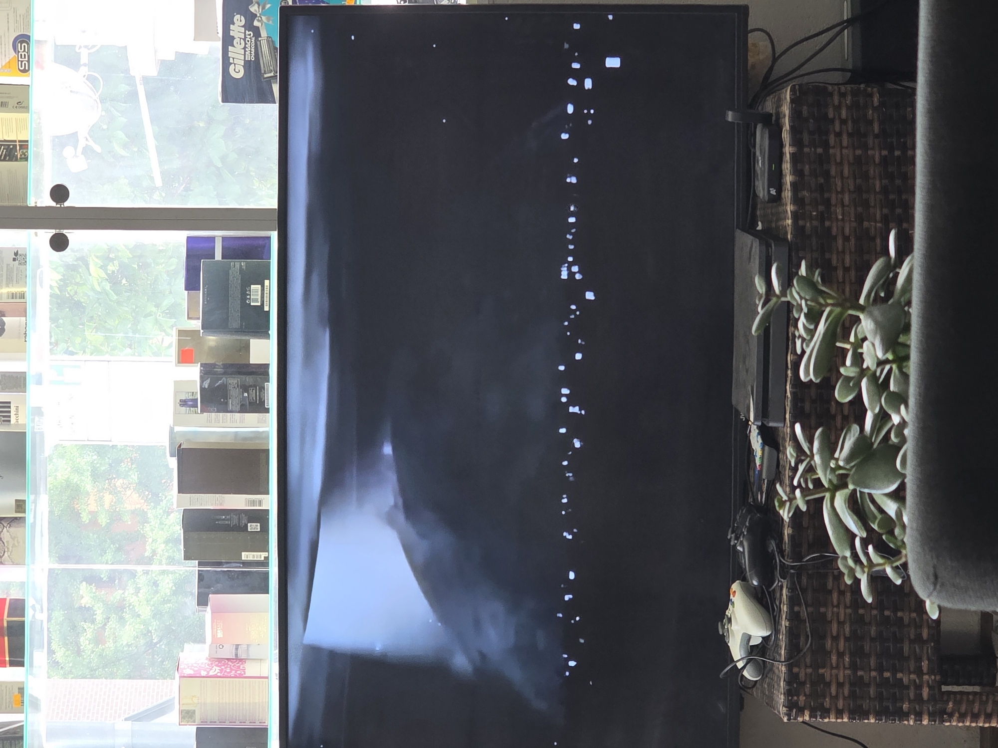 The image shows an indoor scene with a large television screen displaying a dark image with some bright spots. In front of the television, on a woven stand, are two video game controllers, a gaming console, and a small router or set-top box. A succulent plant is in the foreground. In the background, a shelf displays various boxes of products. The setting appears to be a retail environment, possibly a store in Varna, Bulgaria, given the presence of product packaging that might be found in such a location. The lighting is dim, with the television being the primary light source in the foreground.
