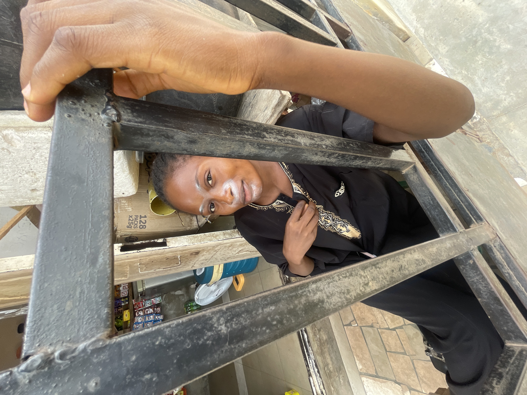 A person is seen looking up through a metal security gate or bars. The person has medium skin tone, dark hair, and is wearing a black garment with gold-colored embroidery. The person has some kind of white substance on their face. One of their hands is placed on the garment. Two hands, also with medium skin tone, are holding onto the gate, one at the top and one at the bottom. The gate appears to be made of black painted metal. Behind the gate, a shop or storage area is visible. There are shelves stocked with various products and boxes. A fan is visible. The location is identifiable as Lokoja, Nigeria because this is mentioned in the prompt. The floor has a tiled pattern. The photo is taken from a low angle, looking up at the person and the gate.