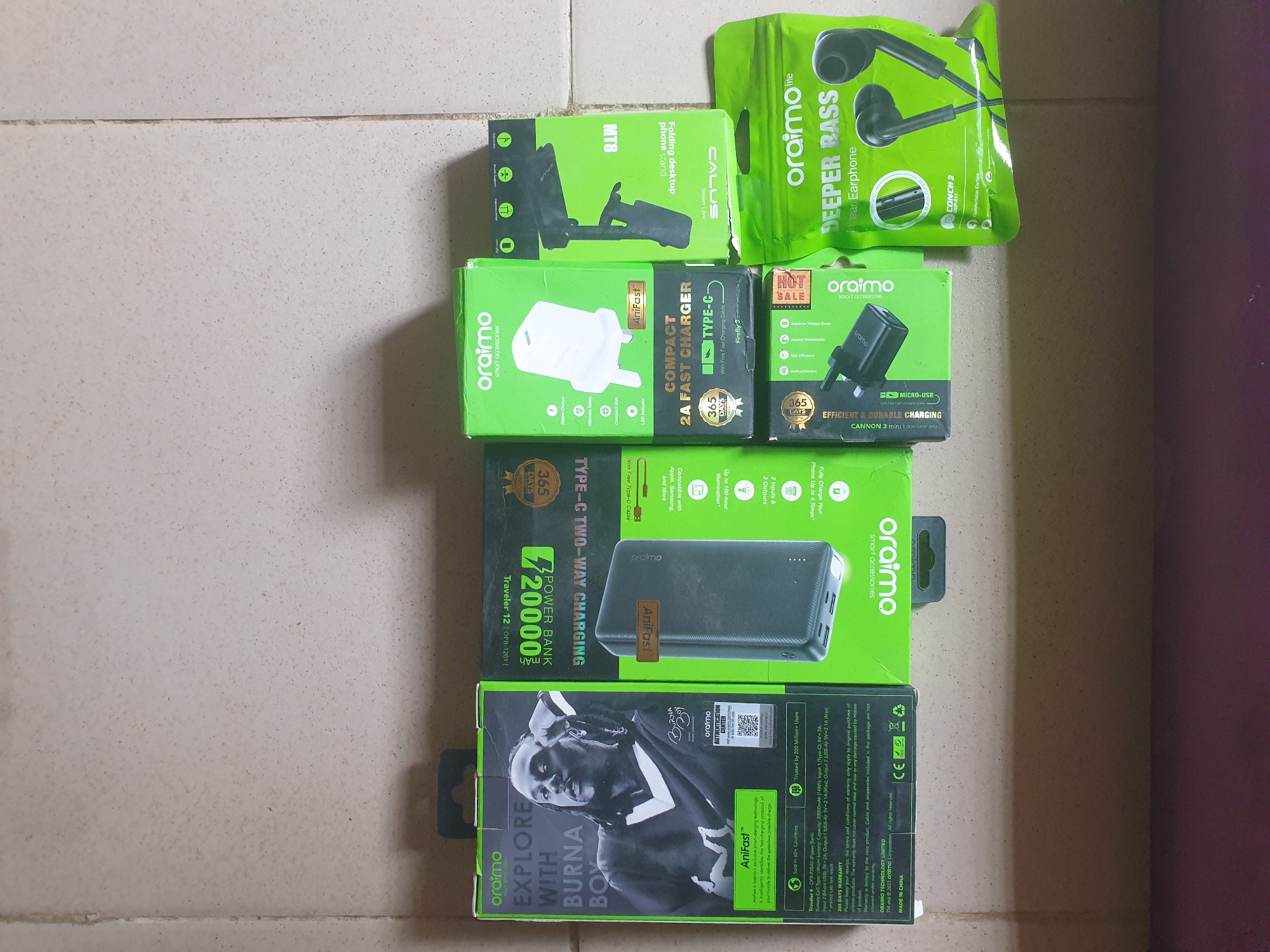 The image shows a collection of oraimo brand electronic accessories, including earphones, a phone charger, a phone stand, and two power banks. The items are arranged on a tiled floor. One of the power banks features a portrait of a person, identified by the text "EXPLORE WITH BURNA BOX," suggesting a collaboration or special edition product. Visible text on the packaging indicates product names like "DEEPER BASS," "COMPACT 2A FAST CHARGER," and "TYPE-C TWO-WAY CHARGING POWER BANK." There are also warranty details and manufacturing information, stating "365 DAYS WARRANTY" and "MADE IN CHINA." The overall scene suggests a retail or personal collection of these electronic accessories. The location context is Anilelerin, Nigeria.