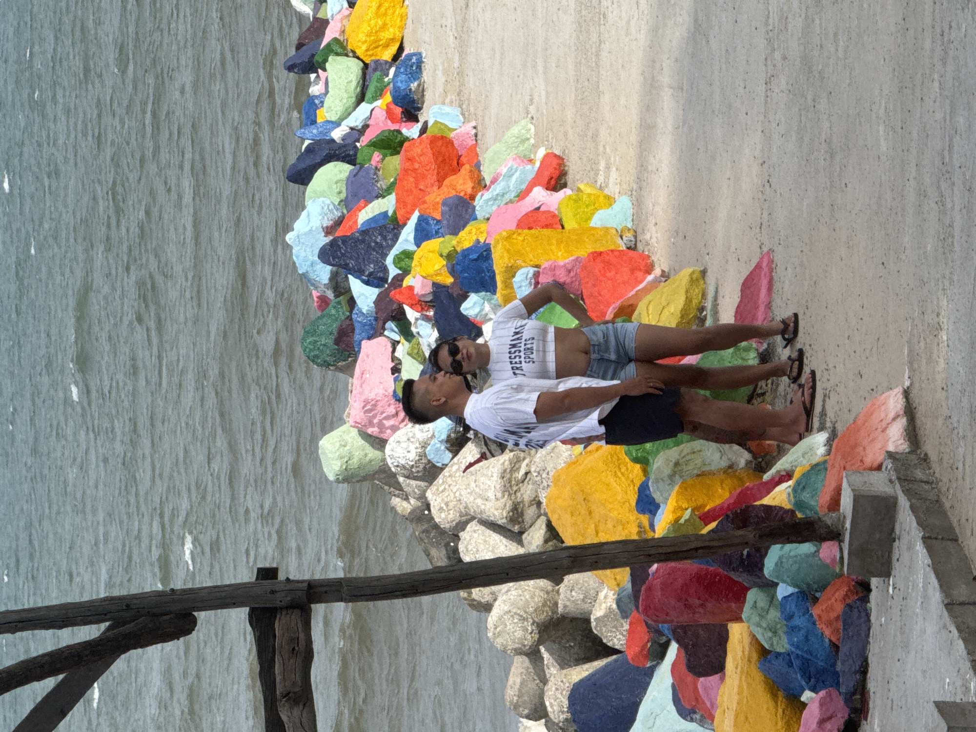 A man and a woman stand on a paved path next to a sea wall made of large, brightly painted rocks. The man, on the left, wears a white t-shirt and dark shorts, and is looking to his left. The woman, on the right, wears a white crop top with "TRESSMAKE SPORTS" printed on it and denim shorts, and is looking at the camera. Both are wearing sunglasses. Behind them, the sea is choppy under an overcast sky. The rocks forming the sea wall are painted in a variety of colors including red, orange, yellow, pink, blue, green, and purple. A weathered wooden post is visible to the left of the couple, partially obscuring the colorful rocks. This scene is located at the Fairy Stream (Suối Tiên) in Phan Thiết, Vietnam, a popular tourist attraction known for its colorful rock formations and stream.