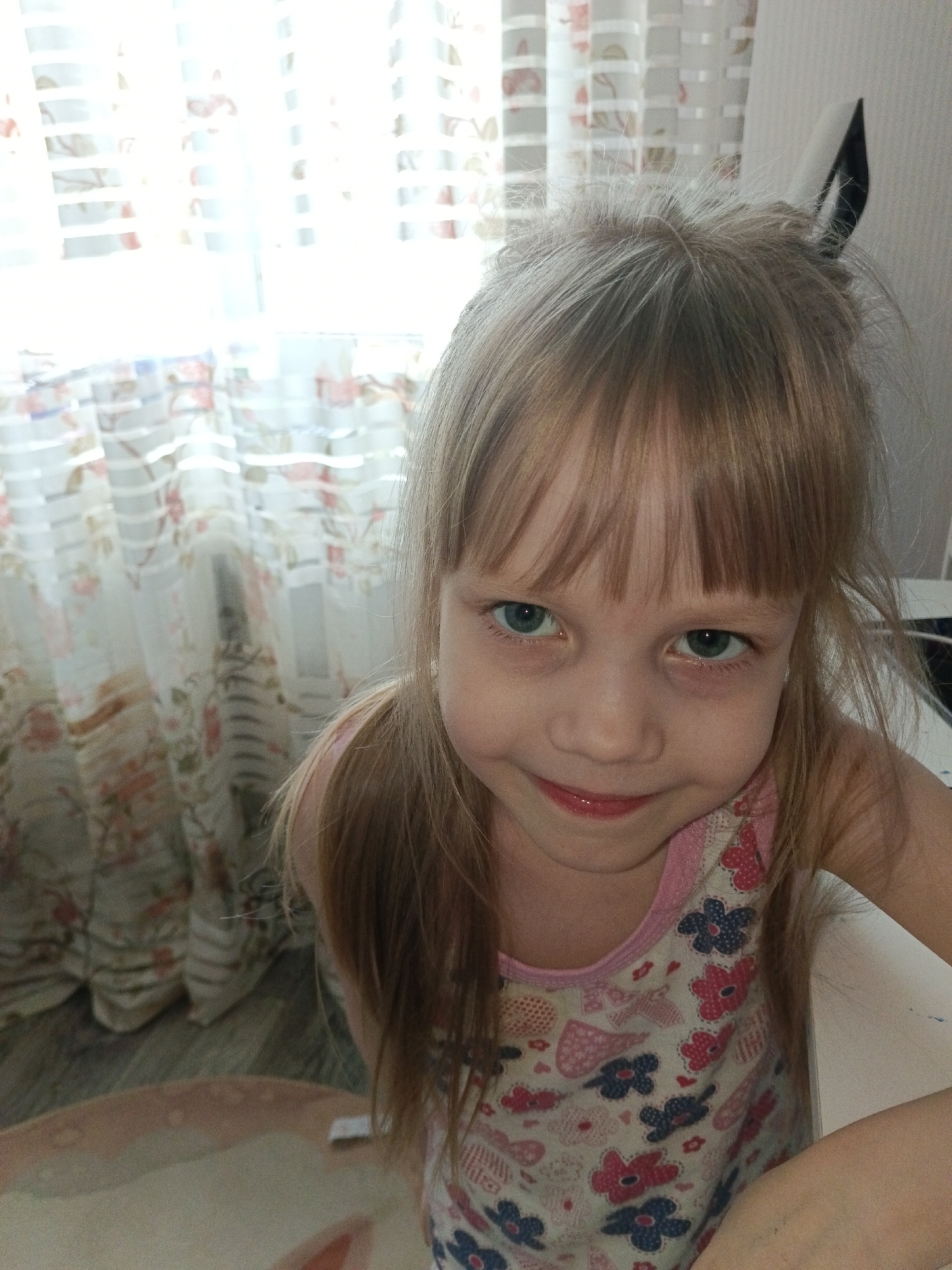 The image captures a young female child in an indoor setting, likely a residential dwelling in Chaikovsky, Russia. The child, approximately 4-7 years old, possesses light blonde or light brown hair with bangs that extend across her forehead, and distinct light-colored (greenish-blue) eyes. She is depicted looking directly towards the camera with a slight, closed-mouth smile. She wears a light-colored tank top with pink trim around the neckline, adorned with a pattern of pink and blue floral elements and other small motifs. Her right arm is partially visible on the right side of the frame, resting on a flat, white surface, possibly a table or desk.
The background primarily consists of a brightly illuminated window on the left, covered by sheer, light-colored curtains featuring a subtle floral design with pink or red blooms and horizontal bands. To the right of the child's head, a plain, light-colored wall is visible. A small, dark, pointed object is observable just above and to the right of the child's head against this wall. The lower-left portion of the frame reveals a section of a light-colored floor, partially covered by a similarly light-toned rug or mat with a faint pattern. The scene is illuminated by natural light entering through the window, resulting in an overexposed area around the window itself.
