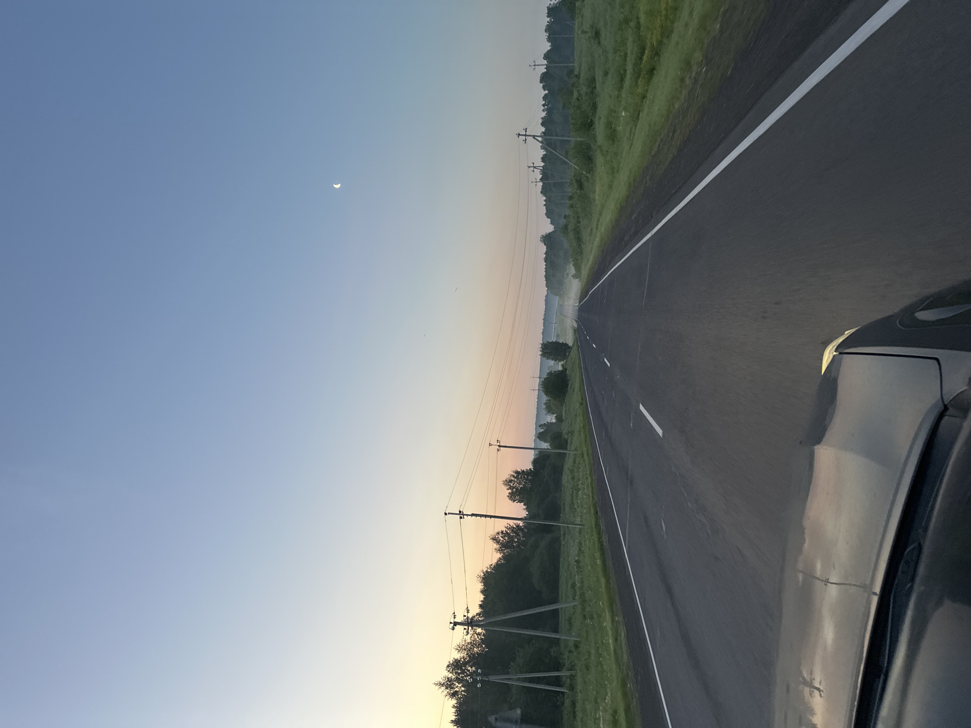 The image shows a view from the driver's seat of a car traveling on a paved road. The road has white dashed lines marking the lanes. The landscape on either side of the road is rural, with green grass and trees. There are utility poles with power lines running along the road. In the distance, there is some fog or mist. The sky is a clear, light blue, with a crescent moon visible. The sun appears to be rising or setting, as evidenced by the soft, warm light on the horizon. The car's hood and windshield wipers are visible in the foreground, reflecting the sky and landscape. The scene is peaceful and serene, suggesting a journey in the early morning or late evening. The location is specified as Krapivinskiy, Russia.