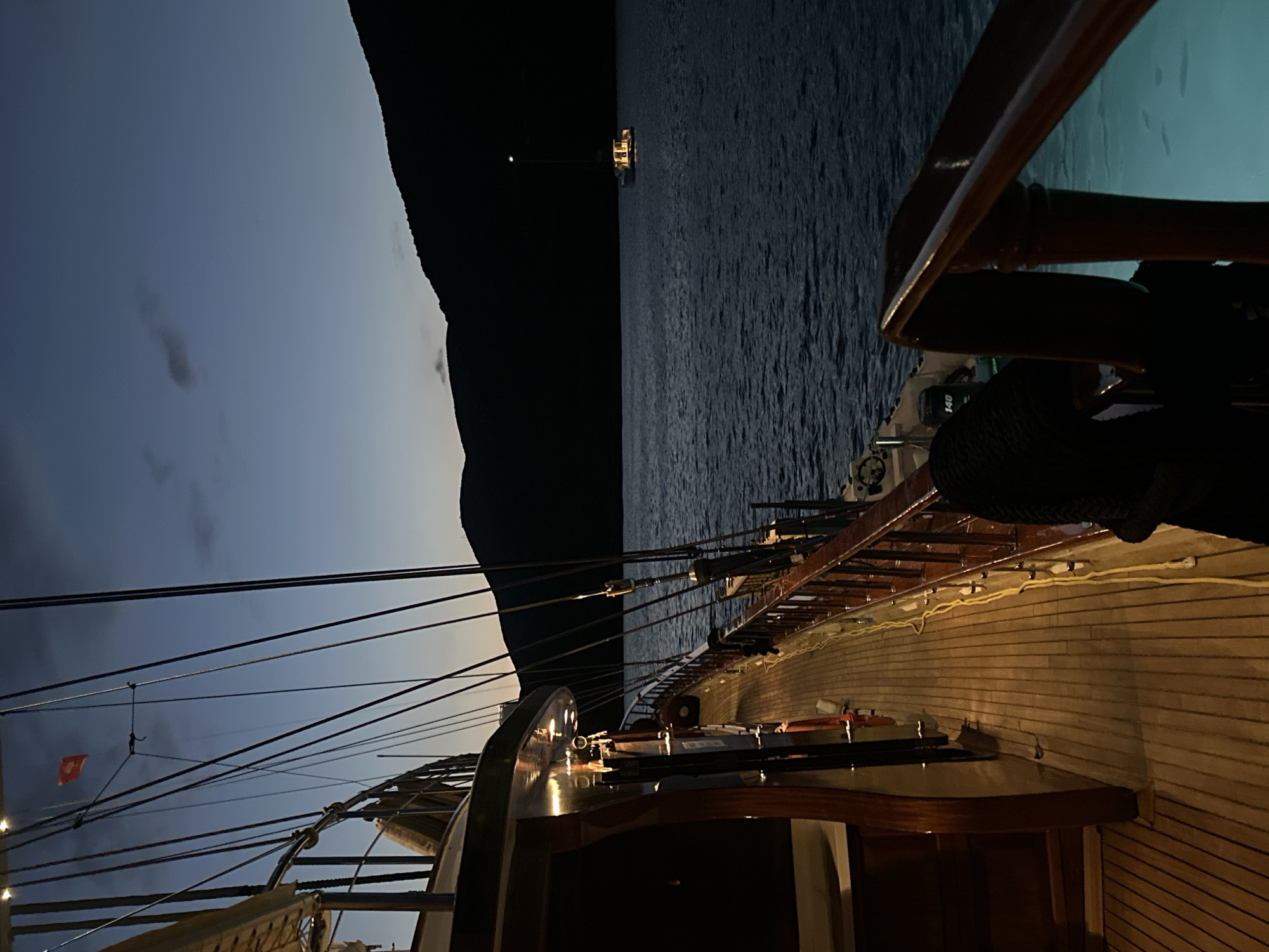 The image, taken at night in Radovići, Montenegro, shows the deck of a wooden sailboat. The deck is illuminated by soft, internal lighting. A dark mountain range is visible on the left side of the frame, silhouetted against a twilight sky. In the distance, there is a lit structure on the water. The sea is relatively calm. The boat's rigging is visible silhouetted against the sky. The overall scene is tranquil and atmospheric.