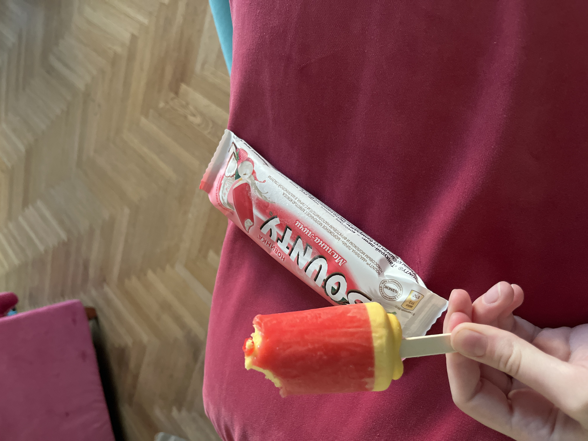Here's a detailed description of the image:
**Objects & Subjects:**
* **Popsicle:** A partially eaten Bounty popsicle with a red (raspberry) exterior and a yellow (lychee) base. The popsicle is held by a hand.
* **Popsicle Wrapper:** A Bounty popsicle wrapper featuring Russian text and graphics.
* **Hand:** A fair-skinned hand holding the popsicle.
* **Background:** A section of a red upholstered seat or cushion. Part of wooden parquet flooring is also visible.
**Actions & Interactions:**
* A hand is holding and presumably eating the popsicle.
**Environment:**
* The setting appears to be indoors, possibly in a home.
* The color scheme is dominated by red, yellow, and light brown tones.
**OCR Details (assumed language: Russian):**
* The text on the wrapper indicates that it is a Bounty popsicle with raspberry and lychee flavors.
* The text also includes nutritional information and other related details.
I cannot infer the location is Moscow, Russia from the image.