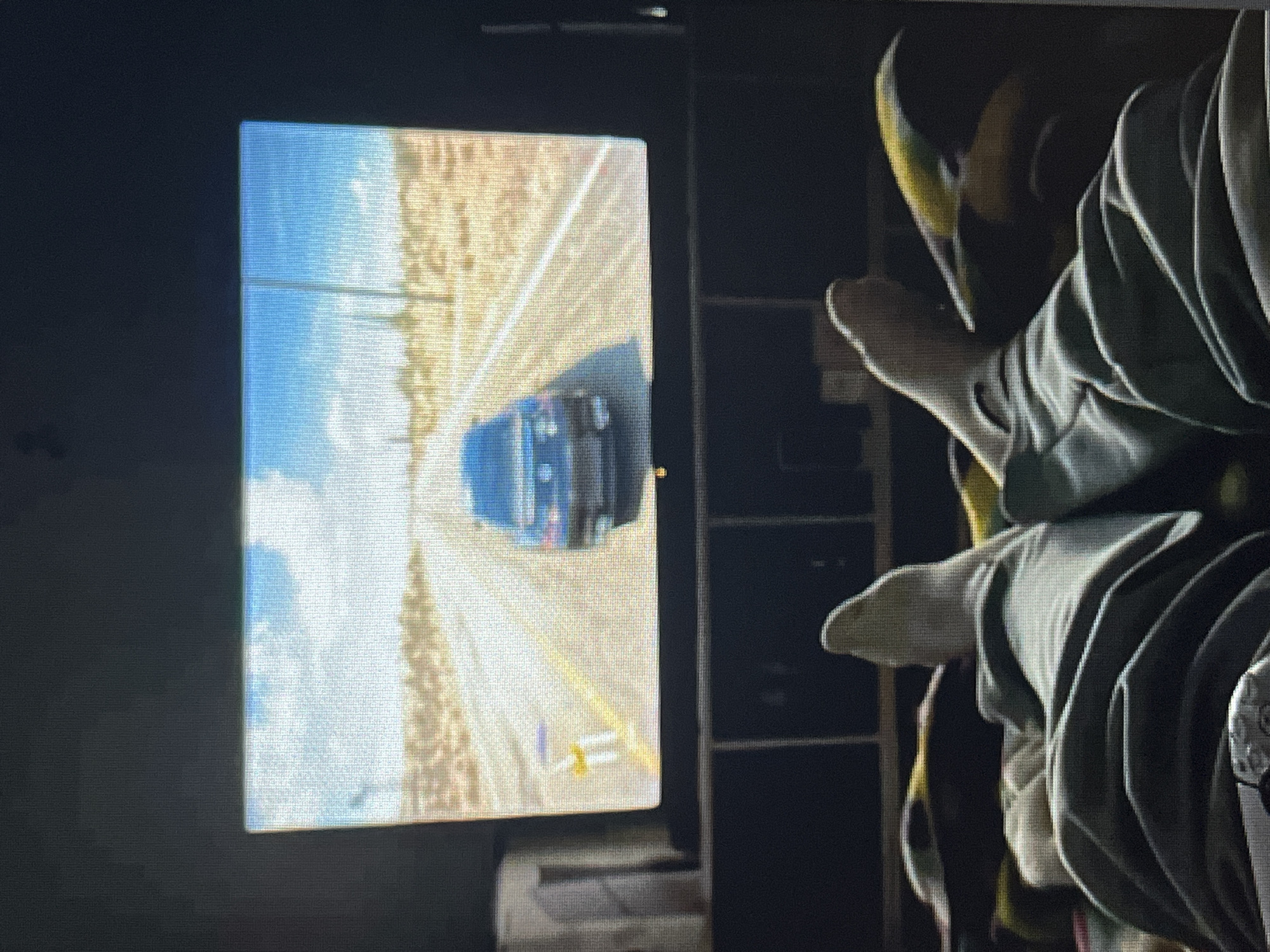 The media file displays an indoor scene where a person is engaged in playing a video game. On the left side of the rotated image, a rectangular screen, likely a television or monitor, is prominently featured. The screen shows a first-person perspective from a video game, depicting a dark-colored car driving on a road. The road appears to be a straight, open stretch, possibly in a rural or desert environment, with light-colored shoulders or dirt bordering it. Yellow road markings and small white or yellow barriers are visible on the side of the road. Above the road, a clear blue sky with white clouds is rendered.
To the right of the screen, a person's lower legs and feet are visible, resting comfortably. The person is wearing light-colored socks and is covered by what appears to be a light green or grey fabric, possibly pants or a blanket, showing visible folds. The feet are slightly elevated, suggesting a relaxed posture. In the darker background behind the person's feet and to the right of the screen, faint outlines of what might be shelves or furniture can be discerned.
The image is captured in a low-light setting, and the screen display exhibits some pixelation and horizontal lines, possibly artifacts from the display itself or the photography process. No discernible text is visible on the screen or in the room. The overall atmosphere suggests a casual and relaxed activity.