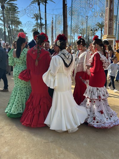 A group of women stand in a public space in Jerez de la Frontera, Spain, likely during a festival. They wear traditional flamenco dresses in colors of green with black polka dots, solid red, solid white with a black fringed shawl, red with a white fringed shawl, and red with a white and red floral patterned bottom. Each has a red flower in her hair. Several men in suits and other dressed people are visible in the background. Palm trees and decorative lights decorate the scene.