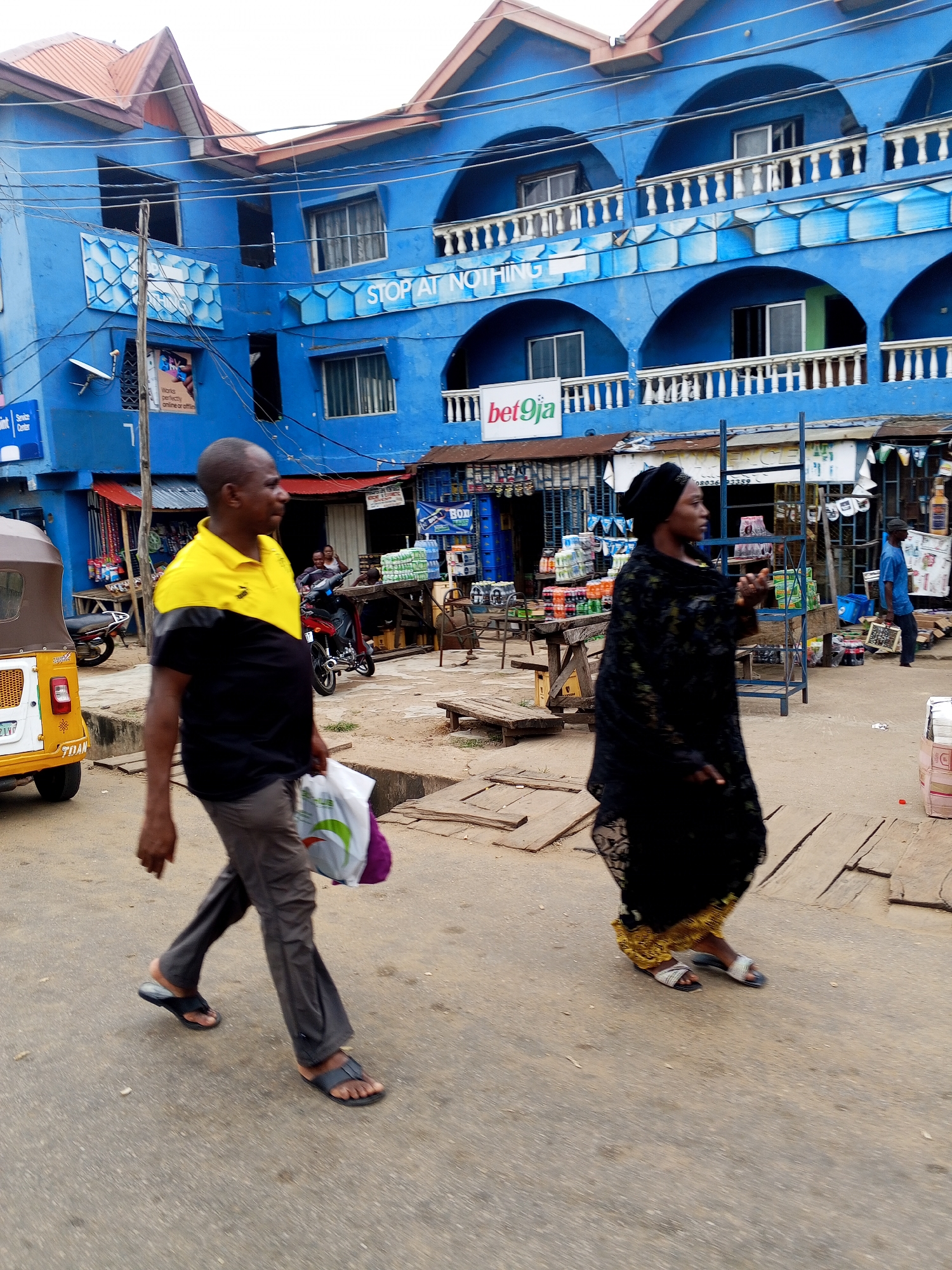 The image depicts a bustling street scene in Lokoja, Nigeria, with a prominent blue building dominating the background. The building features multiple levels with arched windows and balconies, adorned with white railings. Signs advertising "STOP AT NOTHING" and "bet9ja" are visible on the facade. The street level of the building appears to house various shops and stalls selling goods, including bottled drinks and other merchandise.
In the foreground, two individuals are walking from left to right. A man, wearing a yellow and black polo shirt and grey trousers, carries a white plastic bag in his left hand and walks with his right hand by his side. He is wearing black sandals. Beside him, a woman, dressed in a black headscarf and a long black dress with gold trim, walks with her head turned to her right, appearing to be speaking or looking at something. She is wearing silver and white sandals.
To the left of the man, a yellow and brown tricycle, commonly known as a "keke napep" or "okada," is parked on the street. Behind the man, a red motorcycle is visible, with two people seated on it. Further back, another individual in a blue shirt is walking away from the camera, carrying a box. The ground is paved with a mixture of asphalt and wooden planks, suggesting an uneven surface typical of market areas. The overall atmosphere suggests a commercial district with active pedestrian and vehicular traffic.