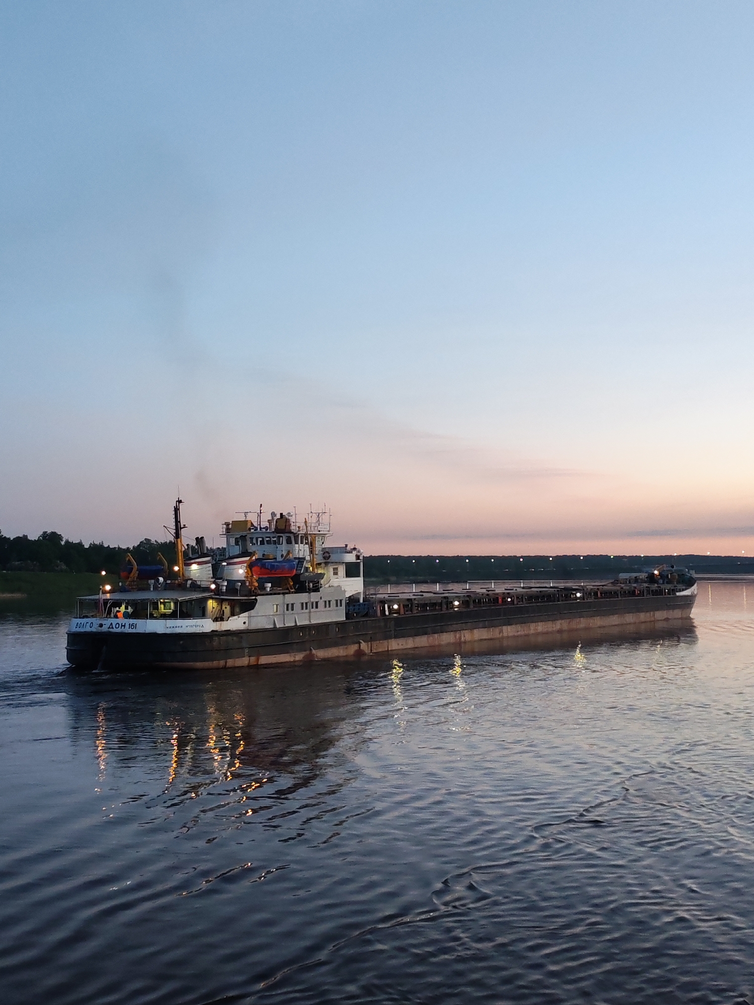 An image depicts a river scene at dusk or dawn in the city of Unknown, Unknown. A large barge, identified as "ВОЛГО - ДОН 161 НИЖНИИ НОВГОРОД," is prominently featured on the river. The barge is partially lit with some lights on. The water reflects the light from the barge. The sky shows a color gradient from blue near the top to pink and orange near the horizon. On the far bank, there is a row of lights, potentially from buildings or a bridge. Some greenery is visible on the near bank.