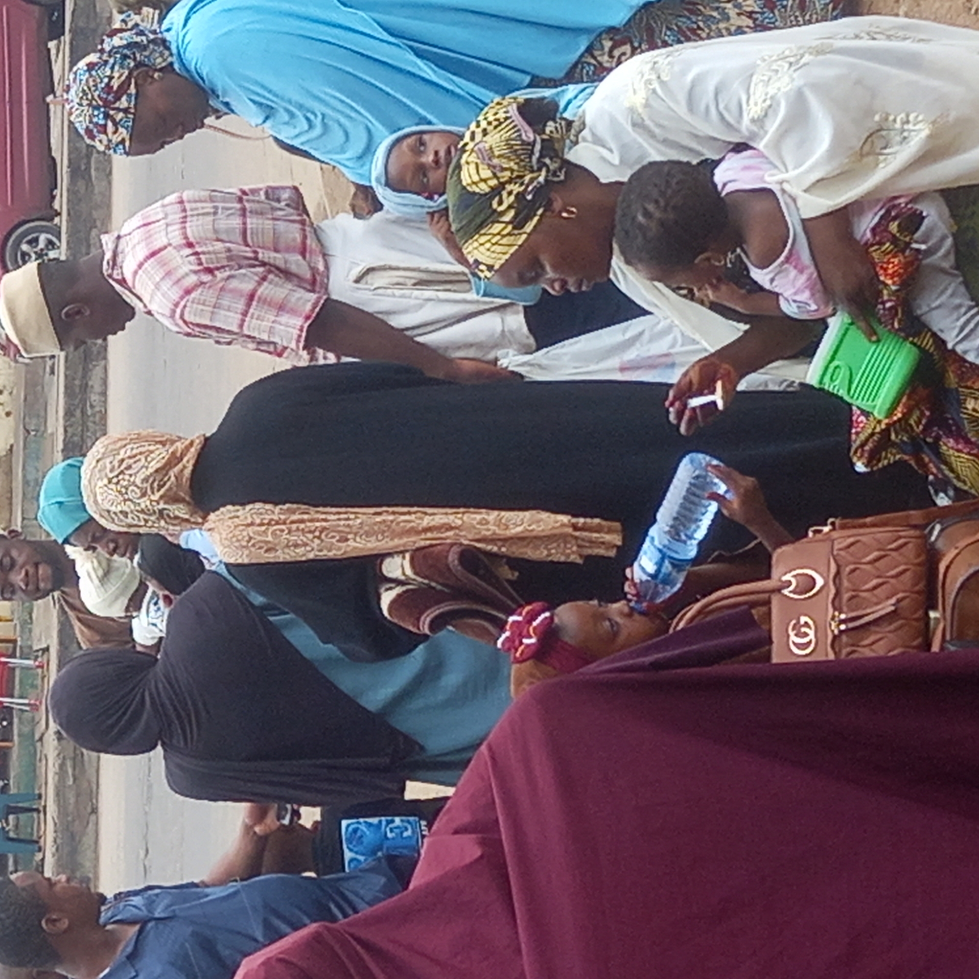In Sardauna, Nigeria, a group of people are gathered outdoors, seemingly attending to a woman lying down. Some of the individuals are offering her water from a plastic bottle. Most of the people are wearing head coverings. A maroon garment and a brown purse with metallic embellishments are in the lower foreground. A parked car is in the background on the left. The scene is on an urban street.