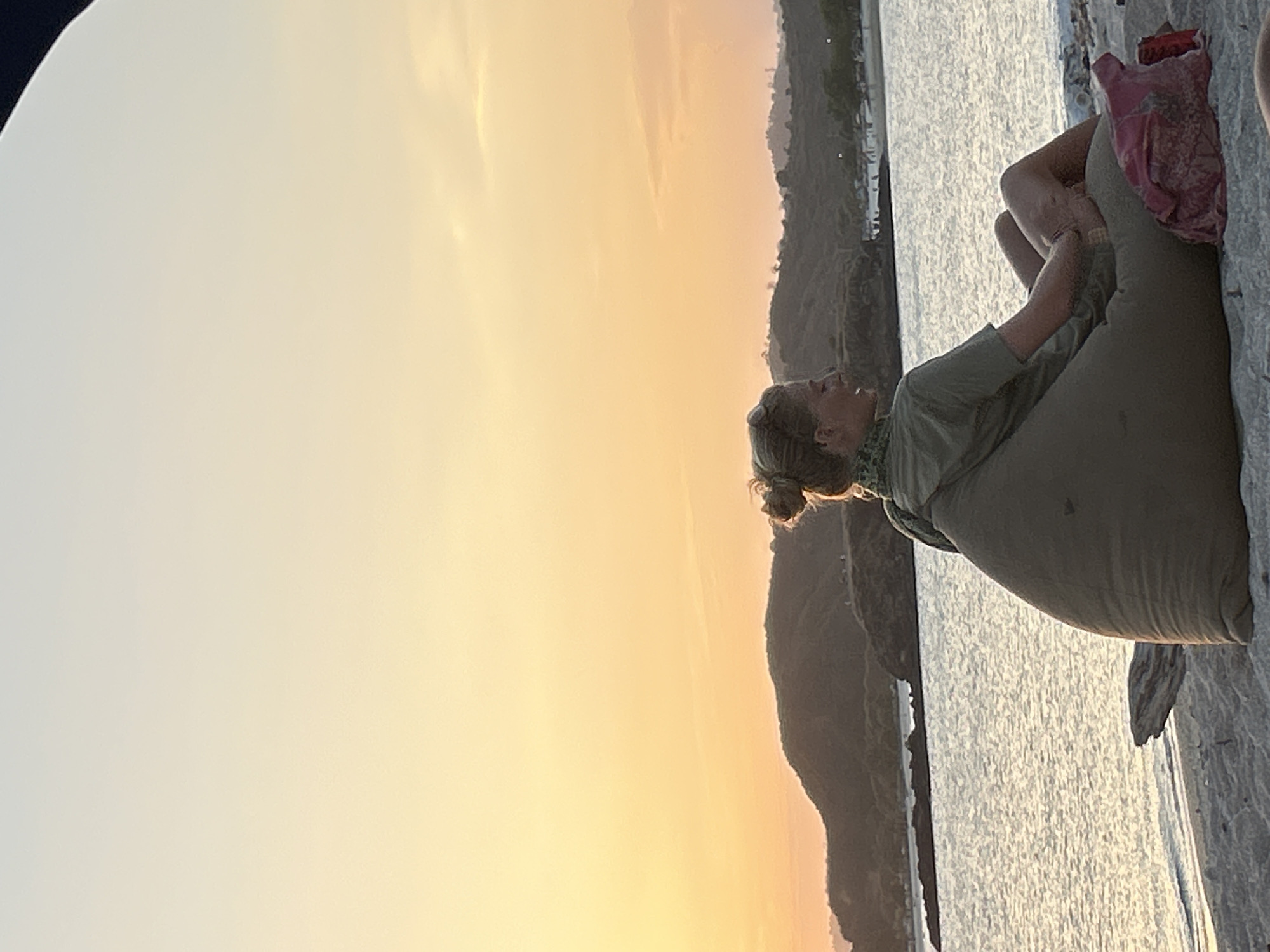 A woman with blonde hair pulled into a bun sits on a large, tan beanbag chair on a sandy beach. She is wearing a green t-shirt and has her legs crossed. In front of her is a pink patterned cloth. The ocean is visible behind her, with gentle waves lapping at the shore. In the distance, there are rolling hills covered in vegetation. The sky above is a gradient of soft orange and yellow, suggesting a sunset. This scene is typical of the beaches in Kuta, Indonesia, known for their beautiful sunsets and relaxed atmosphere.