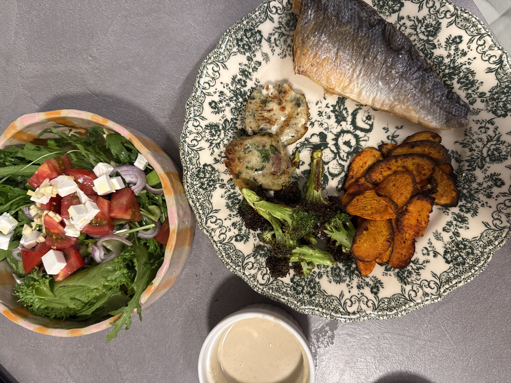 The image shows a meal consisting of a salad, fish, potato cakes, broccoli, and roasted sweet potatoes served on a floral patterned plate. The salad, in a bowl with orange and pink striped rim, appears to have lettuce, arugula, tomatoes, feta cheese, and red onion. The fish is a single fillet, and the potato cakes are round and browned. The broccoli is roasted and slightly charred. A small, white container of sauce is situated near the plate. The meal is placed on a gray surface. The location is Unknown, Unknown.