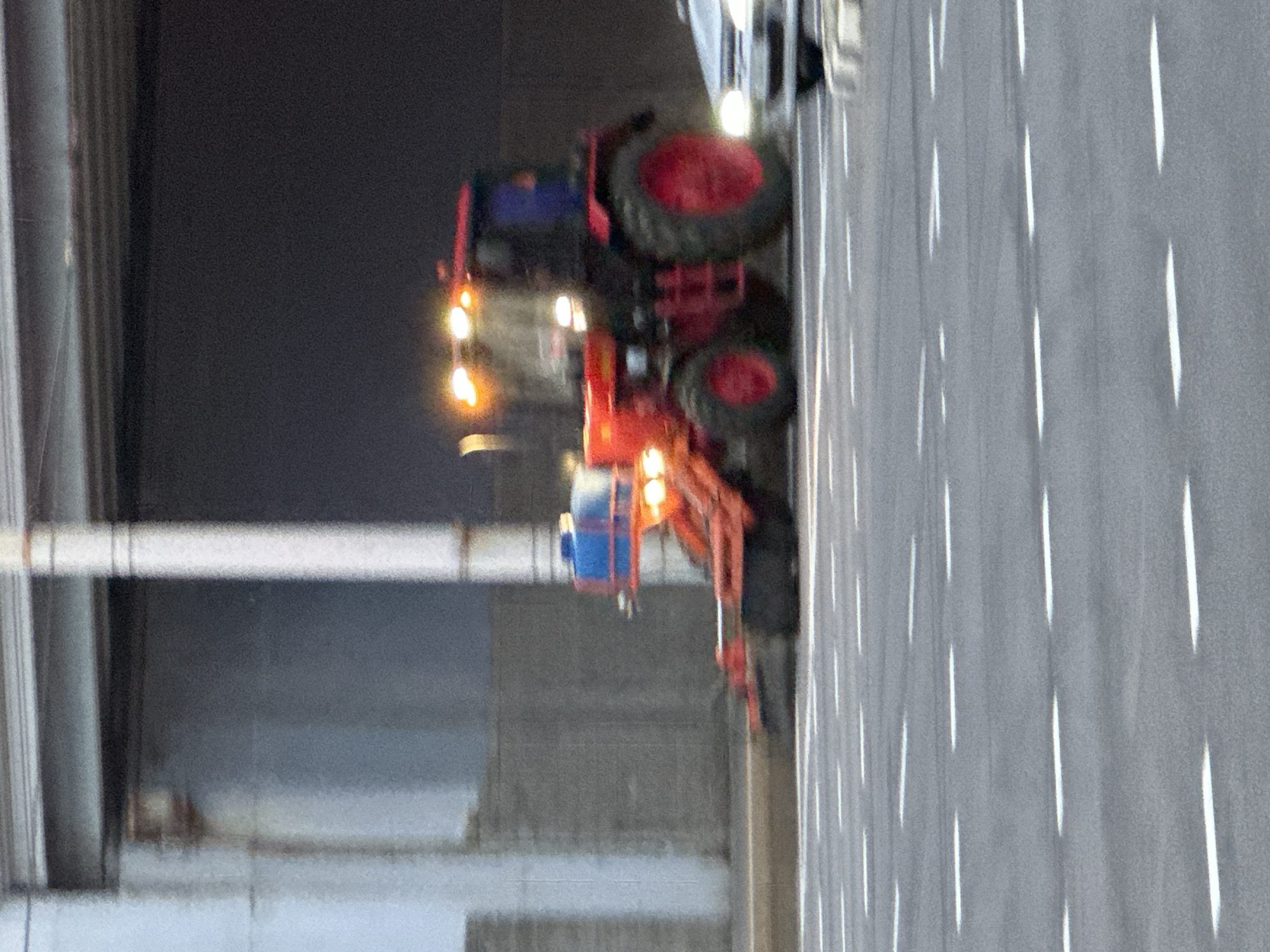 The image depicts a low-light scene on an urban road featuring a red utility tractor equipped with a front-mounted blue tank and an orange attachment, likely a sprayer or sweeping mechanism. The tractor, resembling a Belarus MTZ model, has its headlights and additional work lights illuminated, and its red wheels are visible. It is in motion, exhibiting motion blur. To the right, a light-colored car is partially visible, also with illuminated headlights and exhibiting motion blur. The road features multiple lanes demarcated by dashed white lines. In the background, there is a dark underpass or tunnel entrance, surmounted by a grey structural element with horizontal beams. To the left of the underpass entrance, a light-colored concrete wall or structure is visible, alongside a vertical support column. The overall lighting conditions suggest dusk or night. No individuals are distinctly visible. Based on the prompt's context, the scene is situated within the city of Minsk, Belarus, though no specific landmarks in the image allow for precise geographical pinpointing within the city.