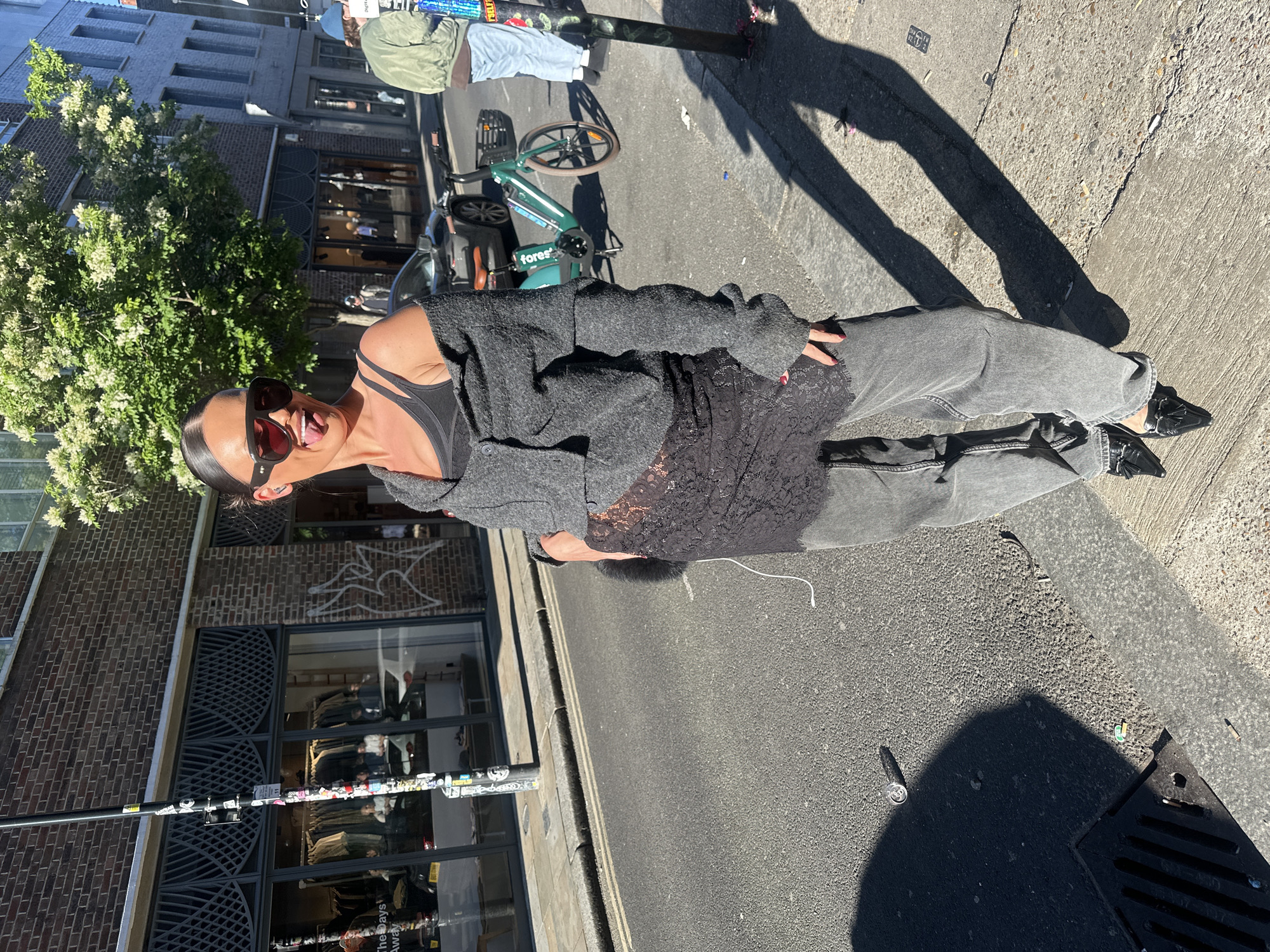 An outdoor, sunlit street scene captures a young woman posing playfully. She is centered in the frame, wearing a dark strappy top, a textured grey button-up cardigan, and washed-out grey jeans. Dark sunglasses cover her eyes, and she is smiling with her tongue slightly out. Her dark hair is pulled back, and she has visible red nail polish. She is wearing black loafers with a decorative buckle.
The setting appears to be an urban street, likely in London as per the context. Behind her to the left is a brick building with several windows, partially obscured by a lush green tree with white blossoms, suggesting warm weather. Further back and to the right, a teal-colored bicycle with "fores" written on its frame is parked. Another person on a bicycle is partially visible in the background. The street features asphalt pavement and concrete sidewalks. Strong, distinct shadows indicate bright sunshine and a clear sky.
On the ground near her feet are clear shadows, including her own, and some street litter: a crushed silver can (possibly a Guinness can, though text is faint), and other small debris near a metal drain cover. A light pole or lamppost is adorned with various stickers, one of which partially reads "The Days Awa...". A white, stylized bird or dove graffiti is visible on a brick wall in the background. The overall activity is a candid or posed moment captured on a sunny day in a city environment.