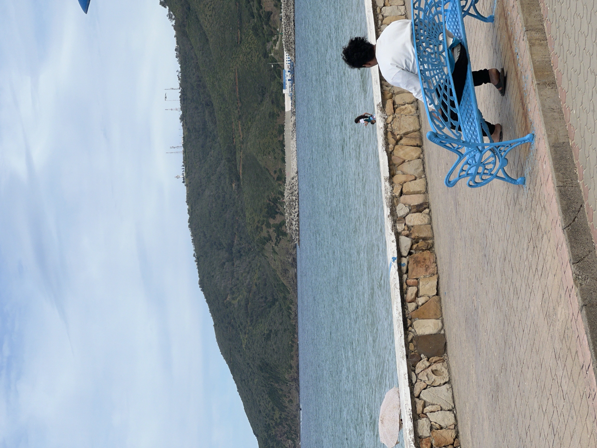 The image shows a scene in M'Diq, Morocco, featuring a man sitting on a blue, ornate metal bench beside the sea. The man is wearing a white shirt and dark pants and appears to be looking down at the water. In the water, there is a person swimming or wading. A stone wall separates the promenade from the water's edge. The promenade is paved with patterned tiles. A green, tree-covered hillside rises up on the opposite side of the water. The sky is partly cloudy.