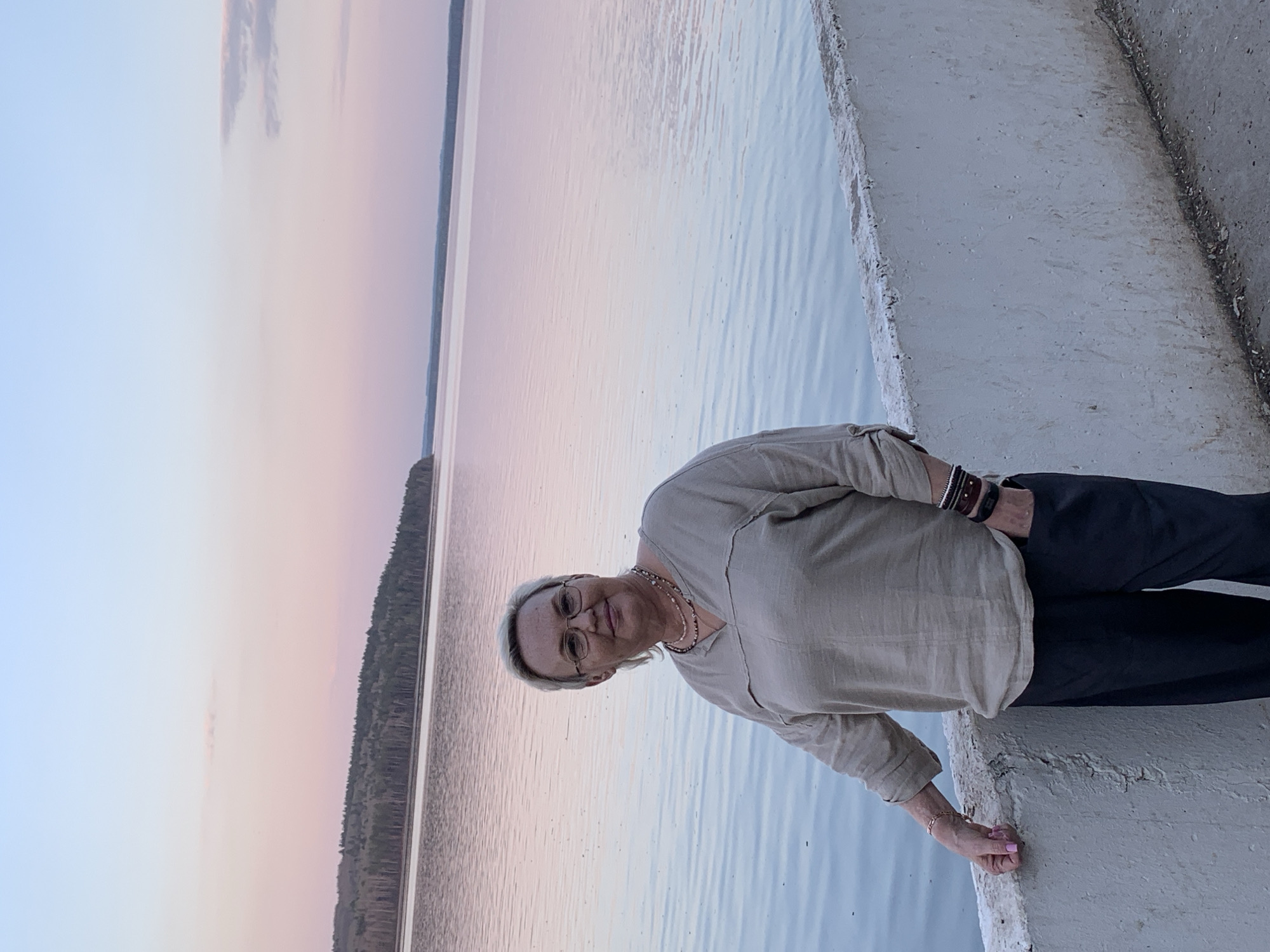 A medium, sideways shot shows a woman standing on a concrete bank next to water with trees visible in the distance. The sky is pink and blue. The woman is wearing glasses, a beige shirt and black pants. She has a necklace and bracelets. Her hand is near the water. The location is likely near a body of water in or near Chaikovsky, Russia.