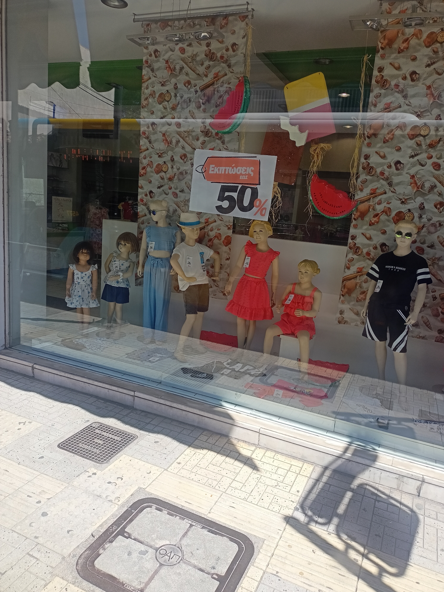 A storefront window displays mannequins dressed in children's clothing, with a prominent sign advertising "ΕΚΠΤΩΣΕΙΣ ΕΩΣ 50%" (Discounts up to 50%). The window is decorated with cutouts of watermelons and ice cream. On the sidewalk outside, manhole covers and textured paving are visible, with shadows cast by street furniture. The overall scene suggests a retail environment in a sunny, urban setting.