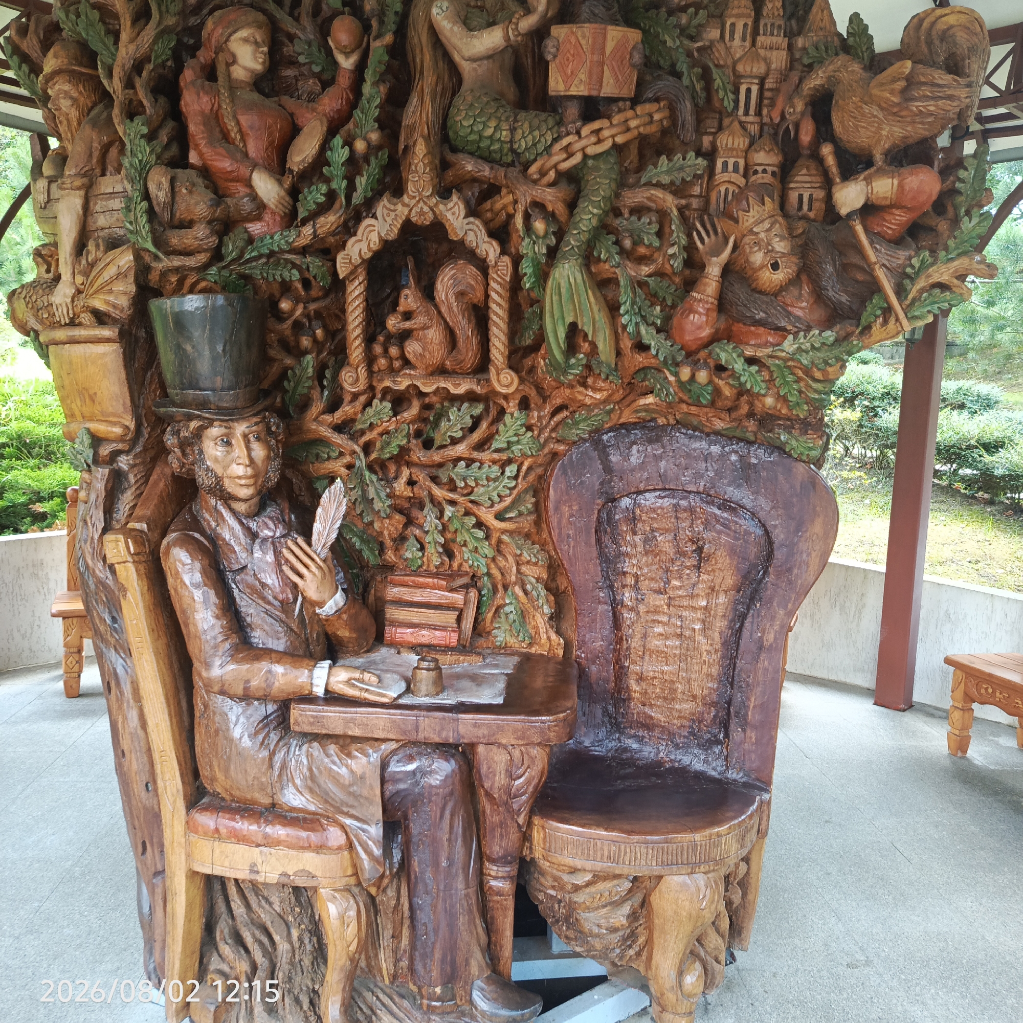 This is a detailed wooden sculpture located in Dagomys, Russia, depicting characters and scenes from Russian folklore. The central figure is a man in a top hat and coat, seated at a table with a quill pen and inkwell, appearing to be writing. Surrounding him are various carved elements including a squirrel in a small arched structure, a mermaid, a woman holding an apple and a mirror, a king-like figure being attacked by a rooster with a hammer, and a representation of a city with onion-domed buildings. The sculpture is intricately carved with organic motifs like branches and leaves, and some parts are painted with subtle colors to highlight details. The sculpture is situated outdoors, under a covered structure with visible support columns, on a paved area with greenery visible in the background. A timestamp is visible in the lower left corner of the image, indicating "2026/08/02 12:15".
