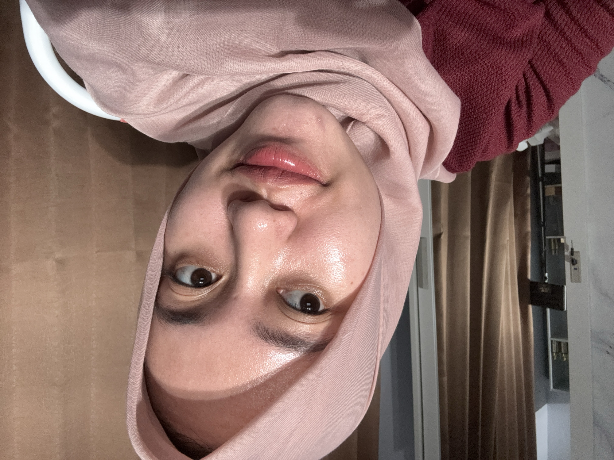 A person wearing a pink hijab and a maroon sweater is photographed indoors. The person has brown eyes and glossy lips. Behind them, there's a white chair and a brown curtain. A door and other household items are visible in the background. The location is Gadingrejo, Indonesia.