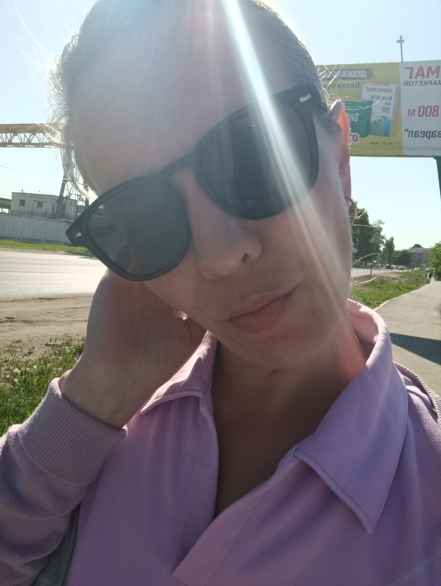 Here is a concise description of the image:
A selfie taken outdoors. The subject is a woman wearing sunglasses and a light purple collared shirt with a sweater, possibly standing on a sidewalk next to a road. In the background, there's a building, a bridge-like structure, and a billboard. The location is likely Chaikovsky, Russia, given the billboard's Russian text. The sunlight is intense and overexposed in some areas.