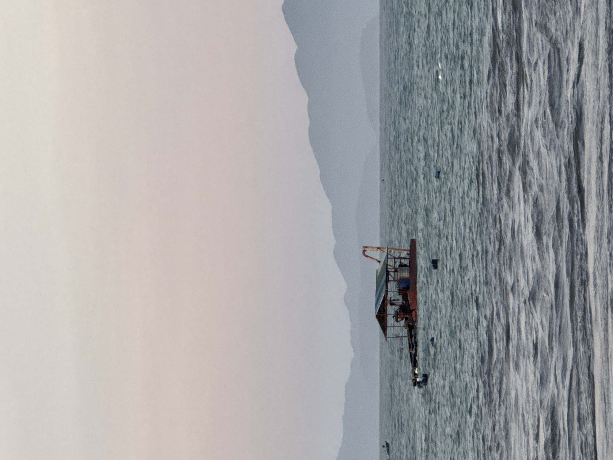 The image depicts a partially cloudy, hazy scene of the Đà Nẵng coastline in Vietnam. The ocean is turbulent, with noticeable wave patterns. A man-made structure, possibly a barge or a small platform, is positioned on the water. Its composition consists of a dark-colored framework and what appears to be a shelter. Mountains are visible in the background, partially obscured by the haze, and a pale sky stretches above. The location of the barge in relation to the coastline suggests it is situated in the sea, not in the immediate vicinity of the shore.