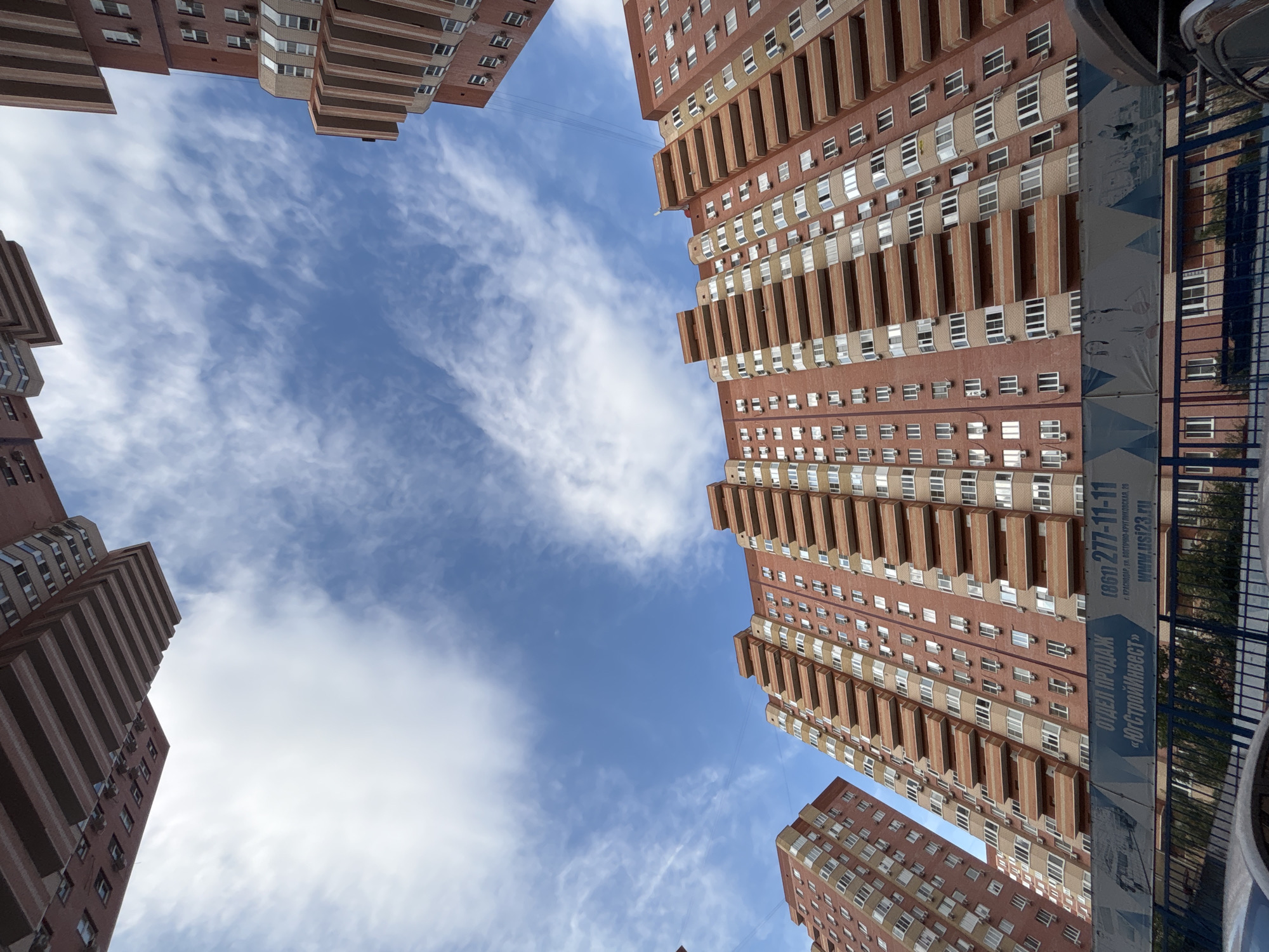 This is an eye-level, upward-facing shot of several tall, brick apartment buildings against a bright blue sky with scattered white clouds. The perspective is wide, looking up from the ground level between the structures, giving a sense of their immense height. The buildings feature repeating patterns of windows and balconies, with prominent horizontal banding that creates a striped effect. Many of the balconies appear to have air conditioning units installed.
On the right side of the frame, a large blue banner with white text is partially visible, attached to a dark fence. The text, which is in Russian, includes a phone number "(861) 277-11-11" and a website "www.usi23.ru". Other visible text identifies "ОТДЕЛ ПРОДАЖ" (Sales Department) and "«ЮгСтройИнвест»" (YugStroyInvest), and a location "Г. КРАСНОДАР, УЛ. ВОСТОЧНО-КРУГЛИКОВСКАЯ, 20" (Krasnodar, Vostochno-Kruglikovskaya Street, 20). This banner, along with the visible text, firmly places the scene in Krasnodar, Russia.
The lighting suggests it is daytime, likely morning or afternoon, given the bright sky and even illumination of the buildings. The weather appears to be clear and pleasant, with the clouds adding visual interest to the sky. There are no people visible in the image. The overall scene depicts a modern urban residential area.